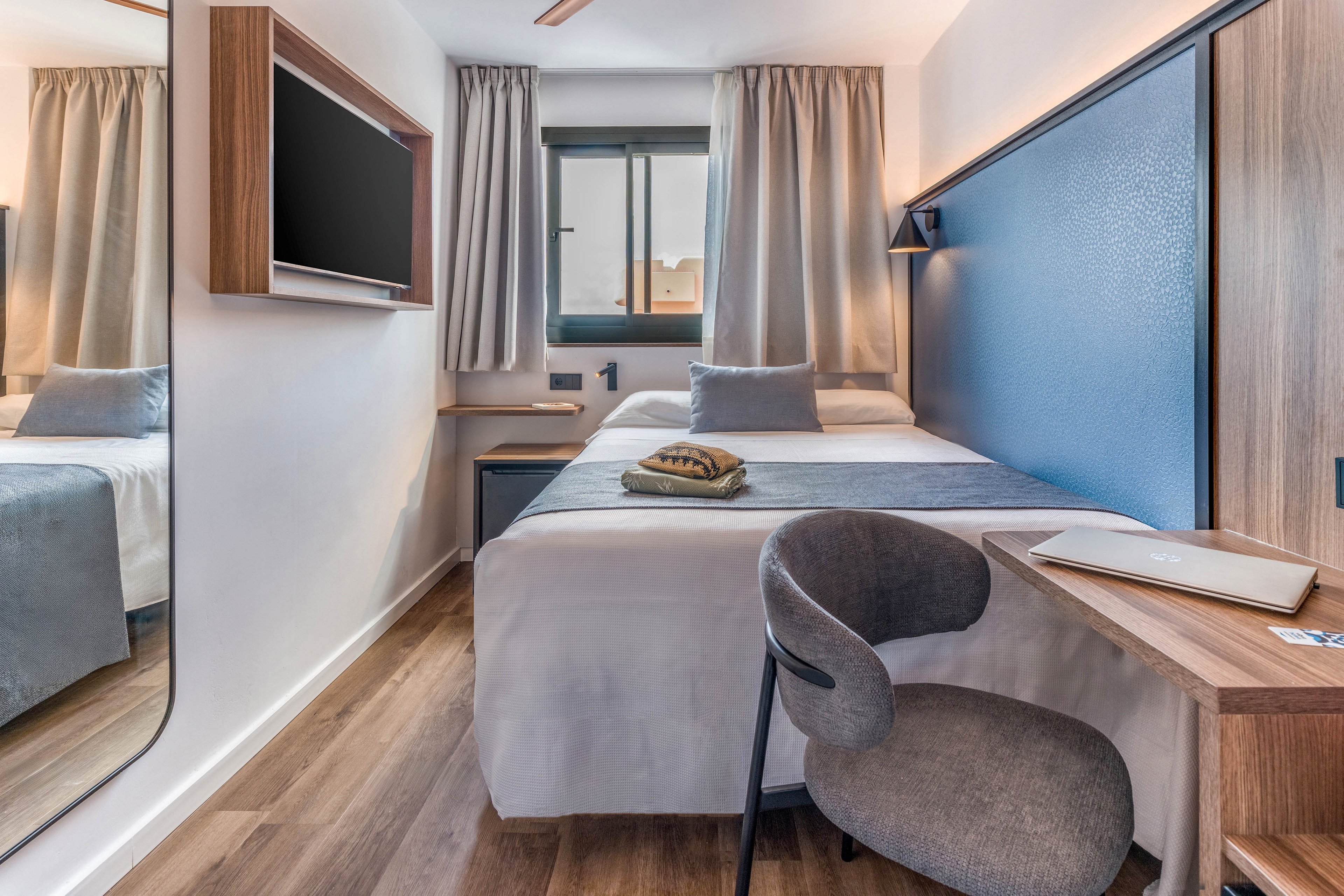 A contemporary compact hotel room features a neatly made single bed with neutral bedding and a blue accent wall. A small wooden desk with a grey upholstered chair sits at the foot of the bed, holding a closed laptop and notepad. A wall-mounted flat-screen television faces the bed, while a large mirror and window with beige curtains enhance the sense of space and light. The room’s clean lines, soft tones, and minimal decor create a calm, functional environment suited for rest or work.