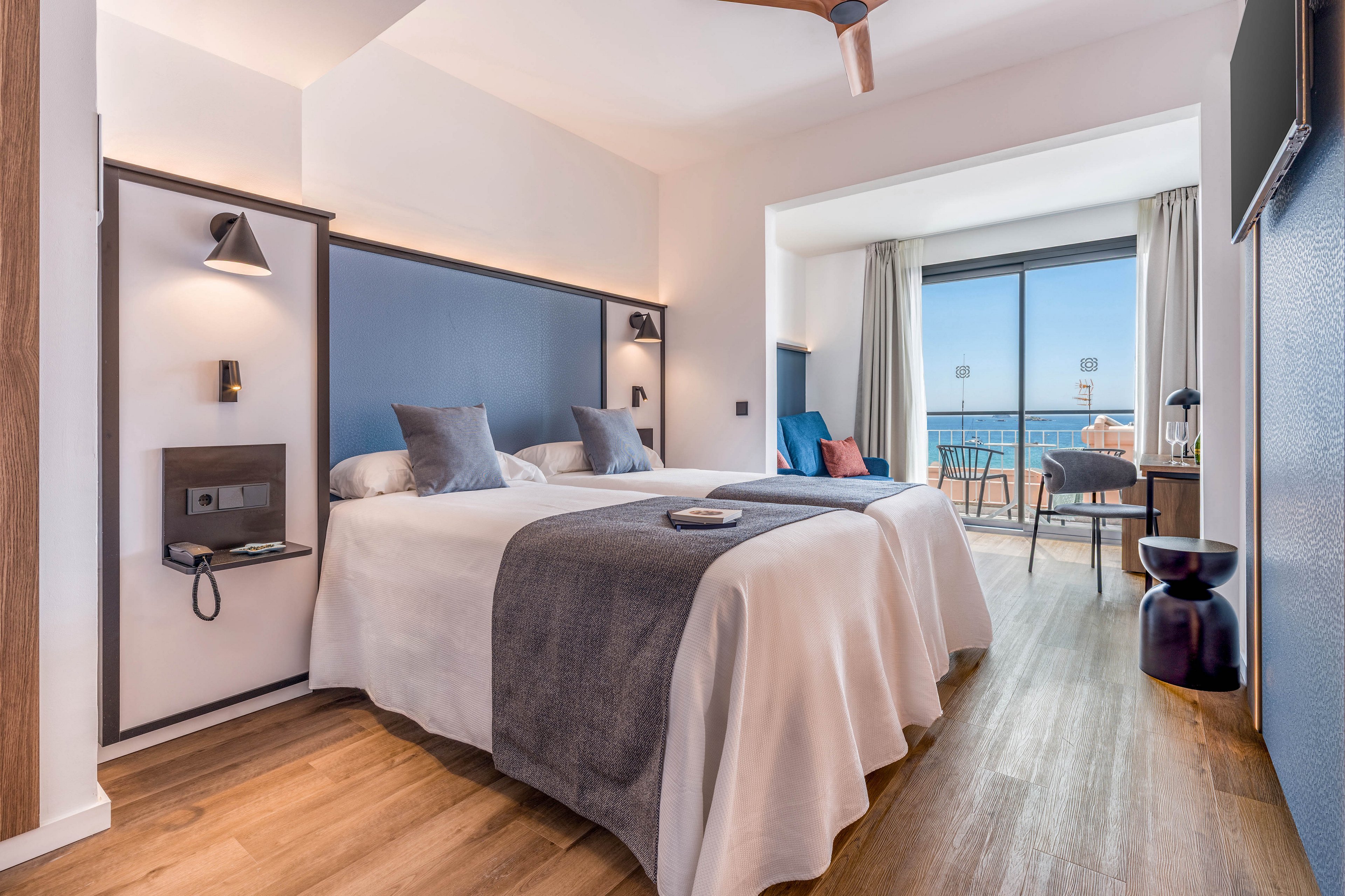 A contemporary hotel room features two neatly made twin beds with white linens and gray throws. The space is decorated in soft neutral tones with blue accent pillows and a minimalist headboard wall. Large floor-to-ceiling windows open onto a balcony overlooking the sea and nearby buildings. Wooden flooring, modern lighting, and simple furniture create a clean, inviting atmosphere.