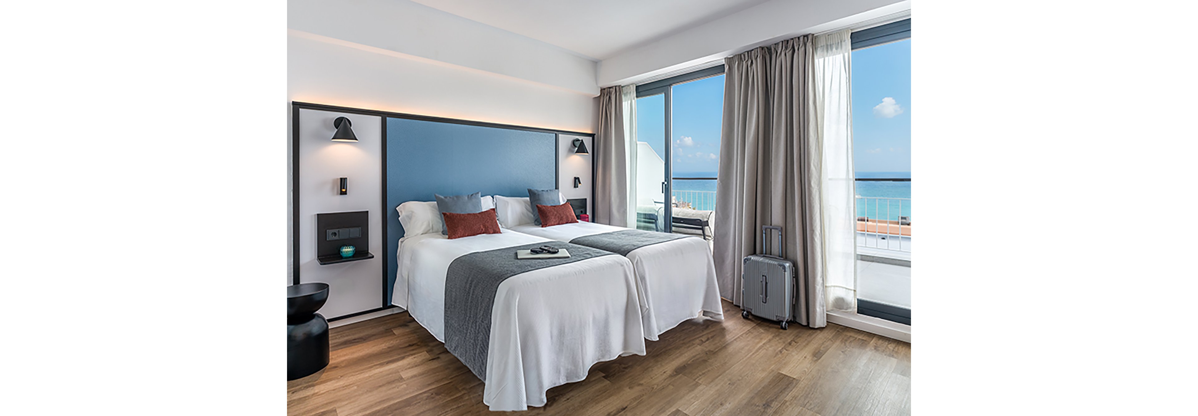 A contemporary hotel room features two neatly made twin beds with white linens, gray throws, and accent pillows. The space includes minimalist wall-mounted lamps, bedside shelves, and a small black side table. Large floor-to-ceiling windows open onto a balcony overlooking the ocean and a clear blue sky. A silver suitcase stands near the curtains, suggesting a travel or vacation setting.