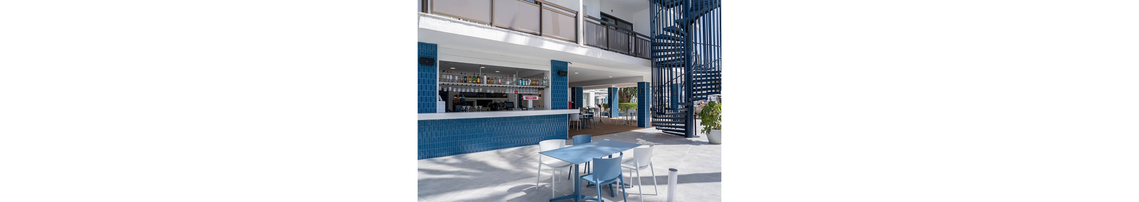 The image shows a contemporary outdoor bar area with a long blue tiled counter and shelves of bottles behind it. In the foreground, light blue and white tables and chairs sit on a spacious patio. A spiral staircase with dark blue railings rises on the right side, adding architectural interest. The setting appears bright and sunny, suggesting a relaxed leisure atmosphere at a resort or hotel.