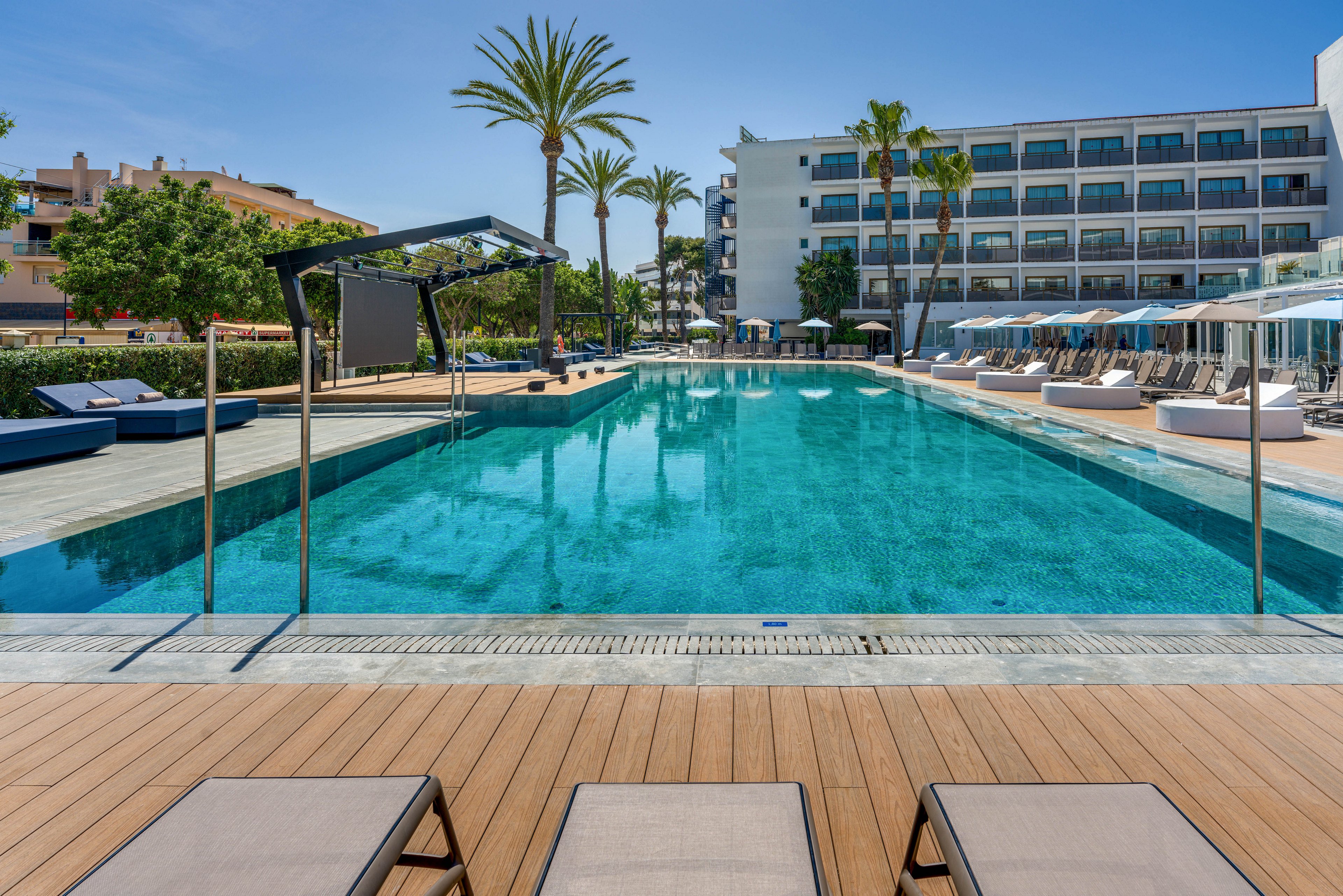 A large, clear swimming pool is the central focus, surrounded by sun loungers and umbrellas. The setting is a contemporary hotel with a multi-story building in the background and palm trees lining the pool area. The scene is bright and sunny, evoking a relaxing and luxurious vacation atmosphere. No visible text or numbers are present in the image.