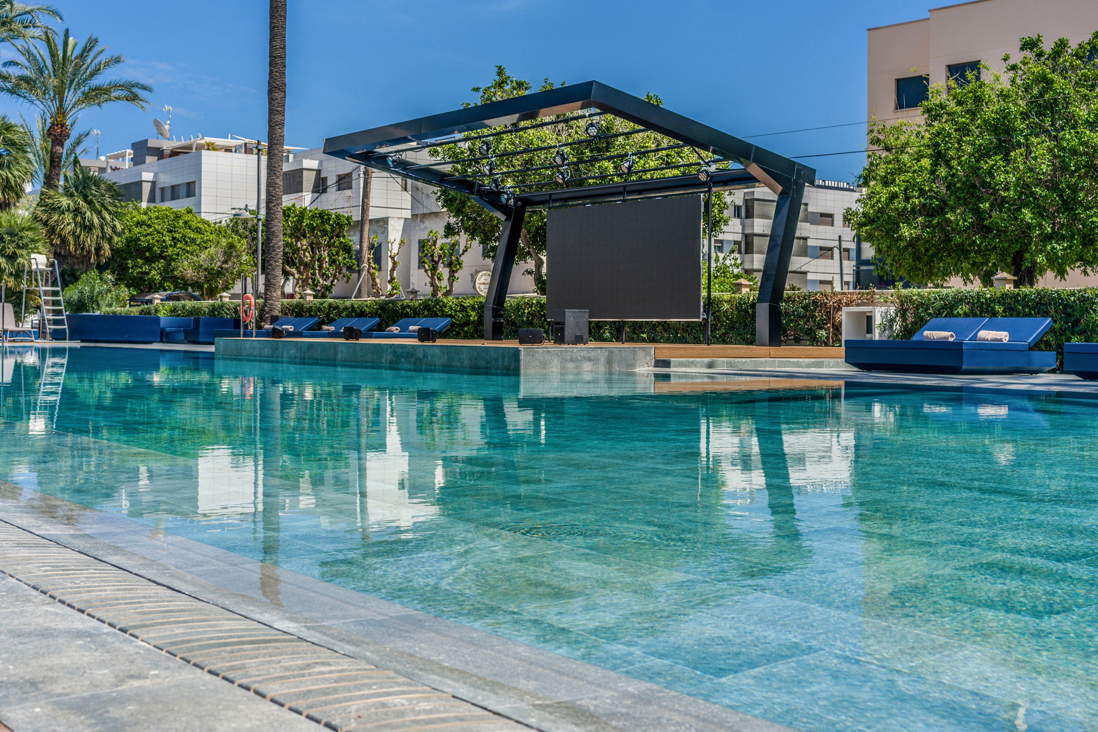 A contemporary swimming pool is featured in the foreground, with crystal-clear water reflecting the blue sky. In the background, a large outdoor stage structure with a screen is visible, surrounded by lush greenery and modern buildings. Several lounge chairs are arranged along the poolside, creating a relaxed and inviting atmosphere.