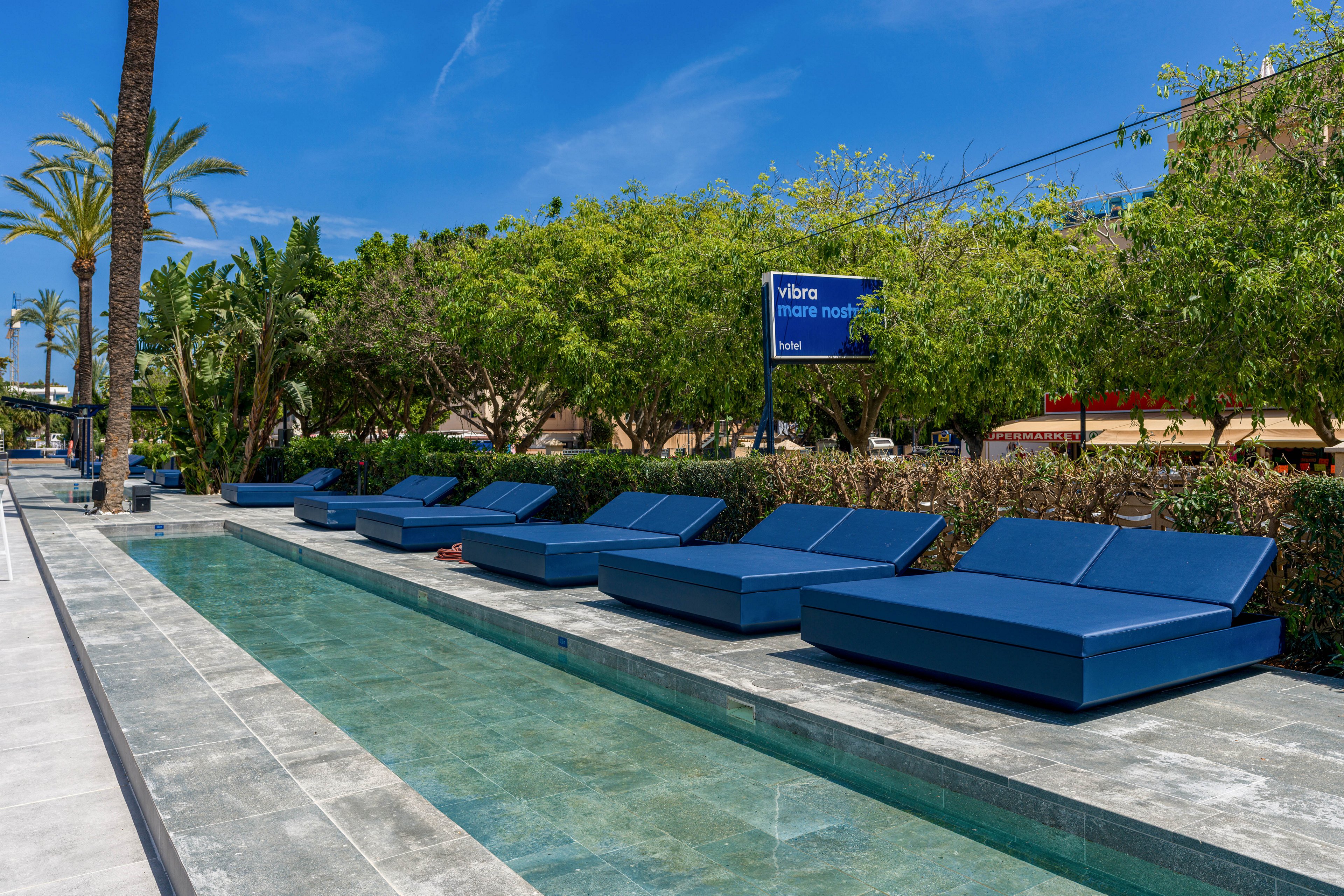 The image features a row of blue cushioned lounge beds arranged along a sleek, shallow pool in an outdoor setting. Lush green trees and palm trees provide a natural backdrop, while a clear blue sky enhances the tranquil mood. A visible sign reads 'Vibra inner hotel,' indicating the location. The scene is calm and inviting, ideal for relaxation in a contemporary environment.