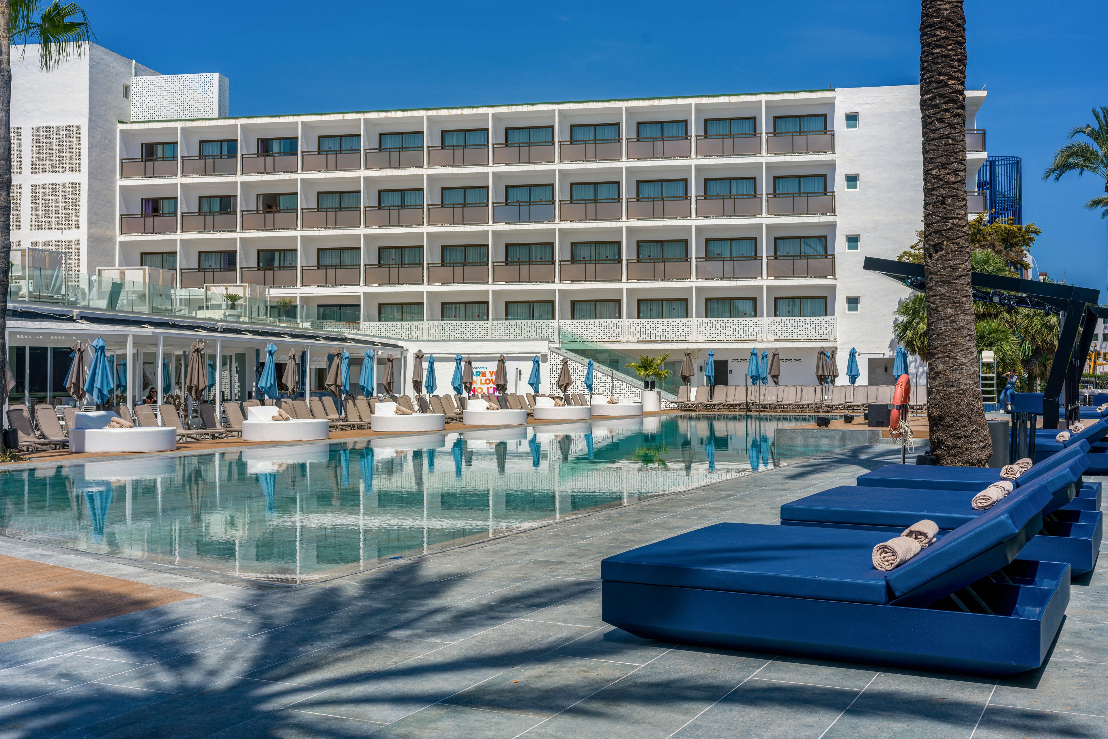 A contemporary hotel building is shown with multiple balconies overlooking a large outdoor swimming pool. Blue cushioned lounge chairs and umbrellas line the poolside, creating a relaxing atmosphere under clear blue skies. The setting appears calm and inviting, with a few people visible in the background. The overall color palette features cool blues and neutral tones, emphasizing a tranquil resort vibe.