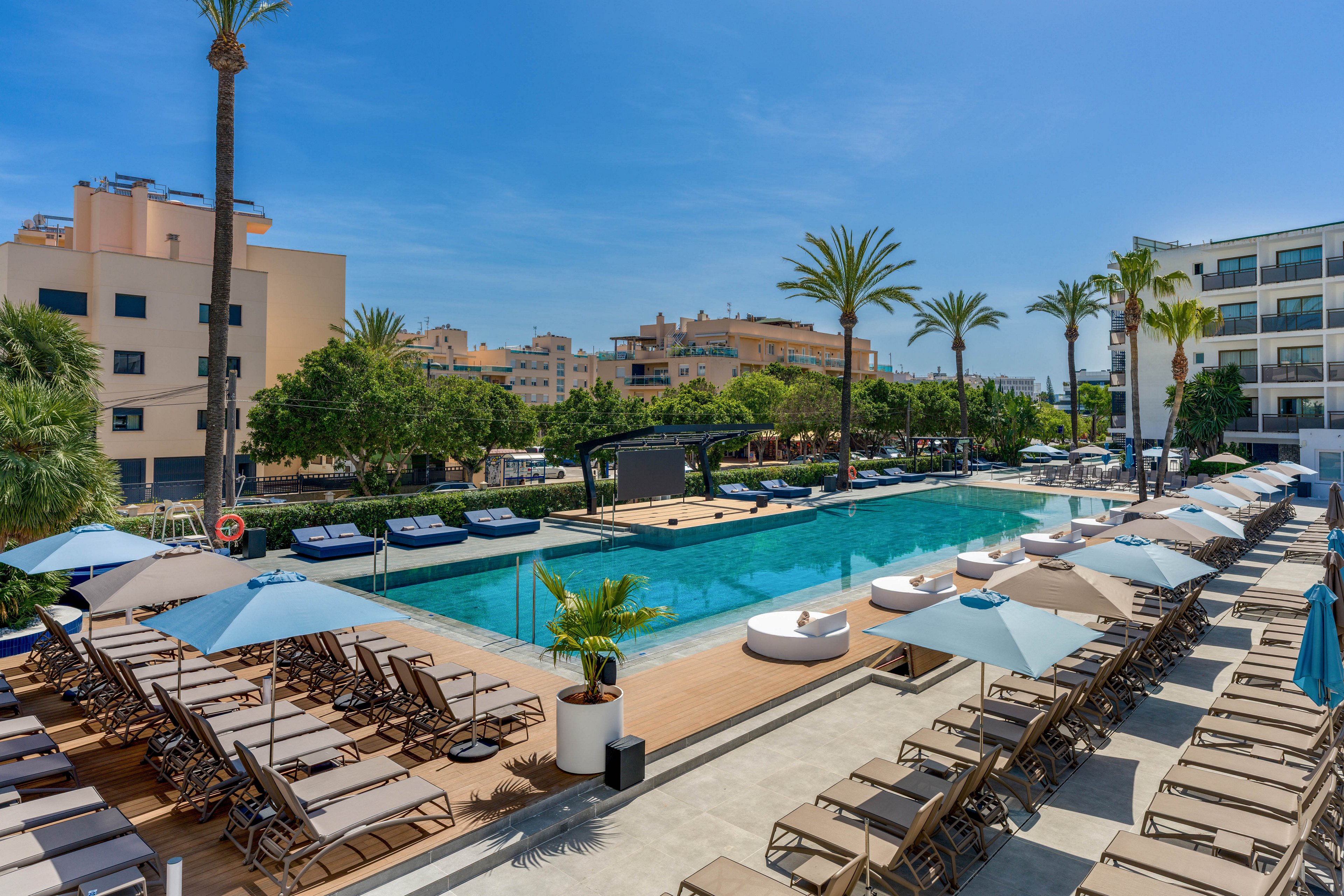 A spacious outdoor hotel pool is surrounded by numerous sun loungers and large umbrellas, set against a backdrop of palm trees and modern buildings. The scene is bright and sunny, with clear blue skies and a relaxed, inviting atmosphere. The pool area appears well-maintained, with clean lines and contemporary design elements. No visible text or numbers are present in the image.