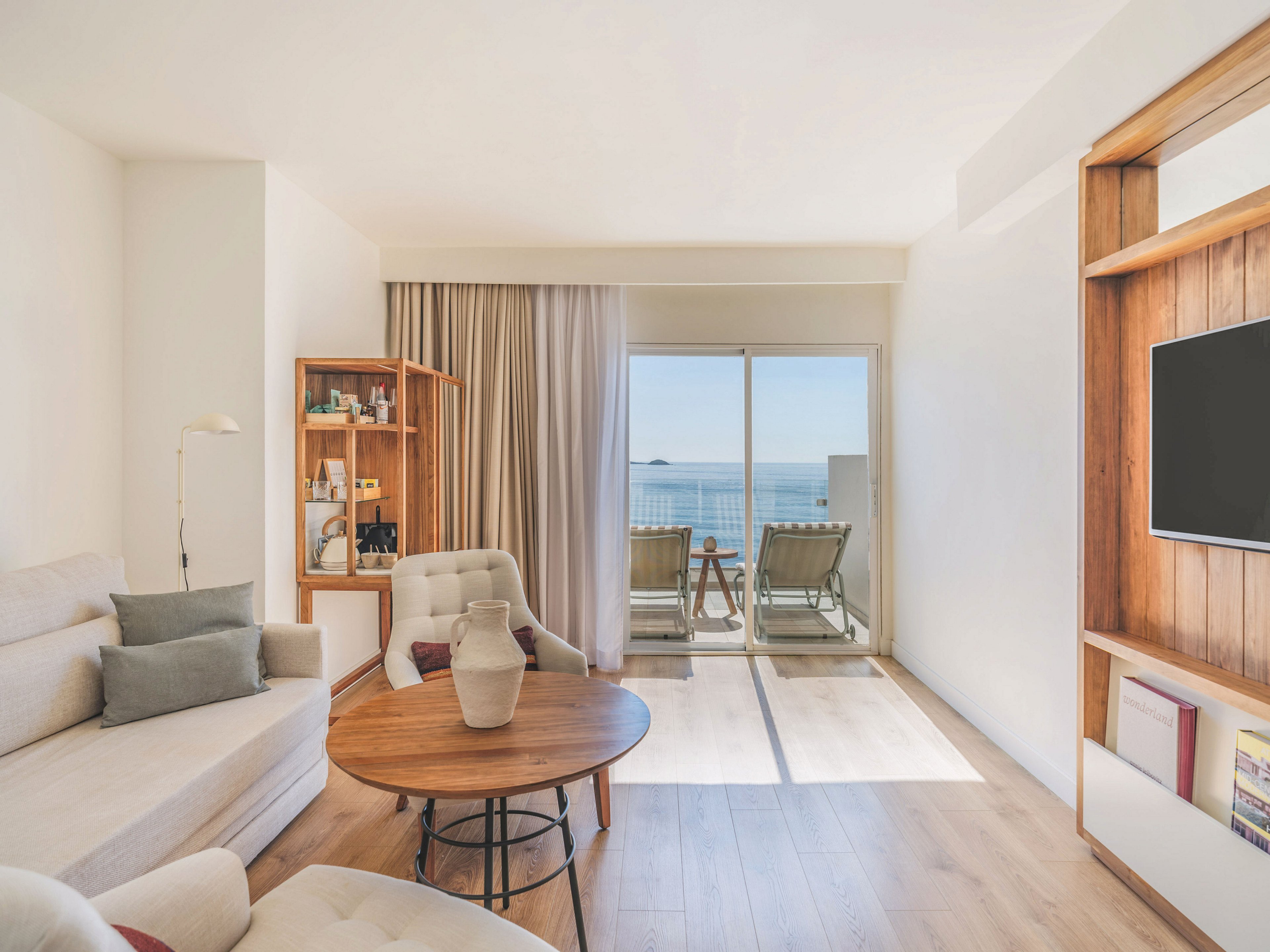 Modern living room with neutral-toned furniture opens onto a balcony overlooking the sea. A light-colored sofa and cushioned chair surround a round wooden coffee table with a decorative vase. Built-in wooden shelving holds a flat-screen television and books, while another shelf unit displays bottles and decor. Floor-to-ceiling curtains frame the glass doors, letting in abundant natural light and highlighting the serene ocean horizon.