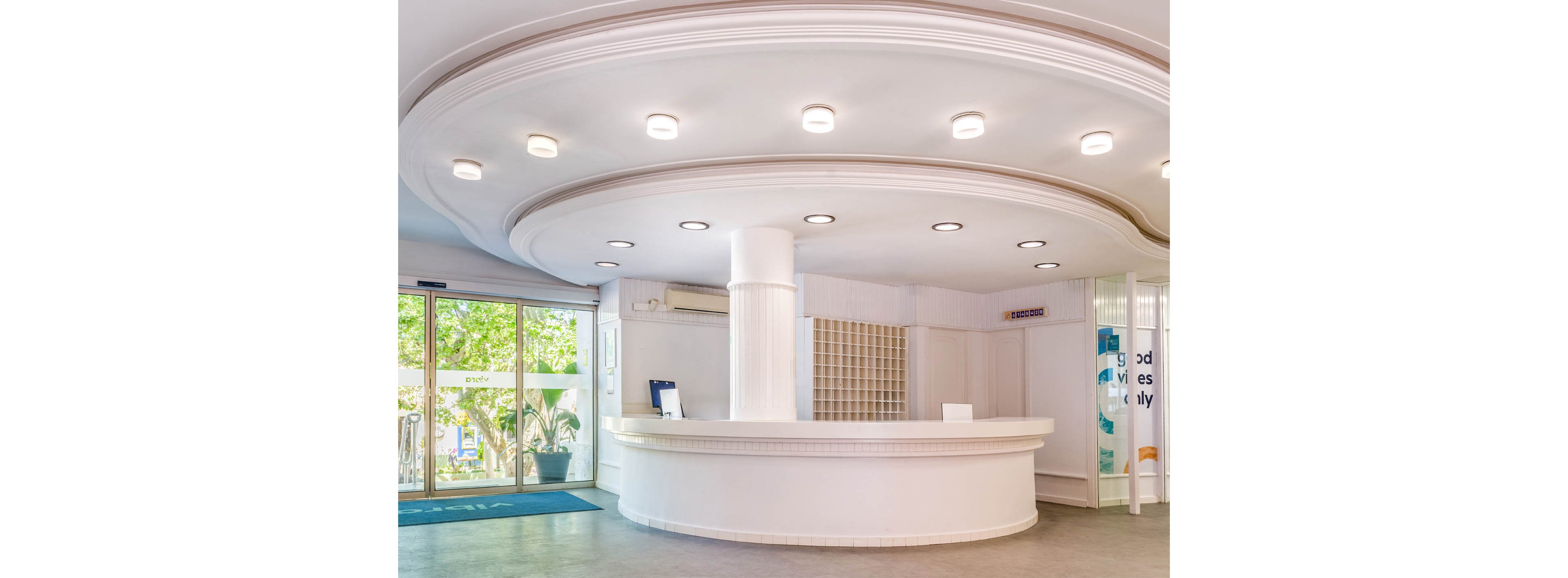 Spacious white reception desk stands in a bright hotel lobby. A curved ceiling with multiple recessed lights illuminates the circular counter below. Glass entrance doors reveal greenery and natural light outside. The scene feels clean, minimal, and welcoming, with no people present.
