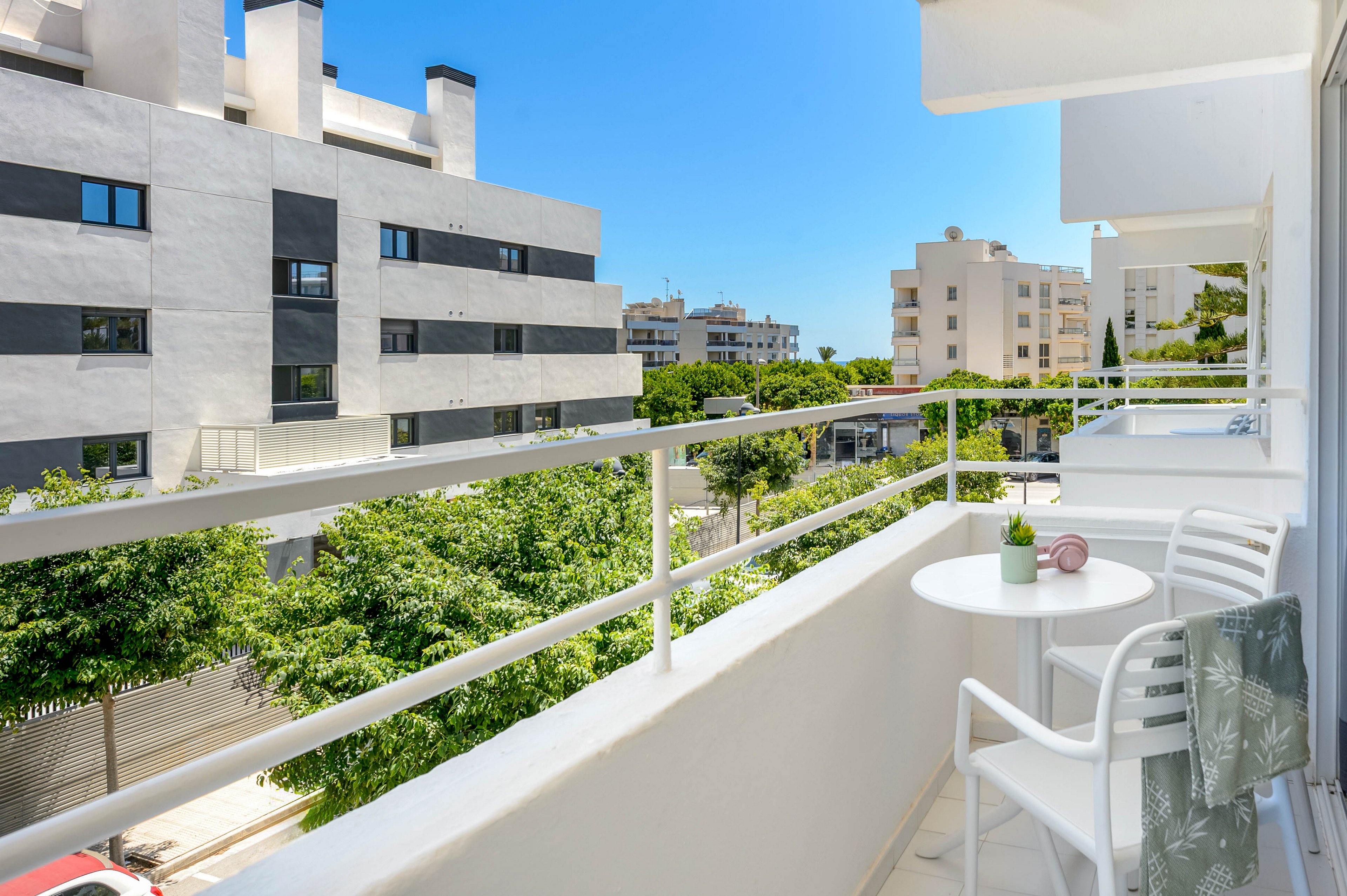 A bright, modern balcony features a small white table and two chairs, overlooking contemporary apartment buildings. The setting is urban and residential, with lush green trees lining the street below. The table holds a small potted plant and a decorative item, while a towel is draped over one chair. The sky is clear and blue, creating a fresh and inviting atmosphere.