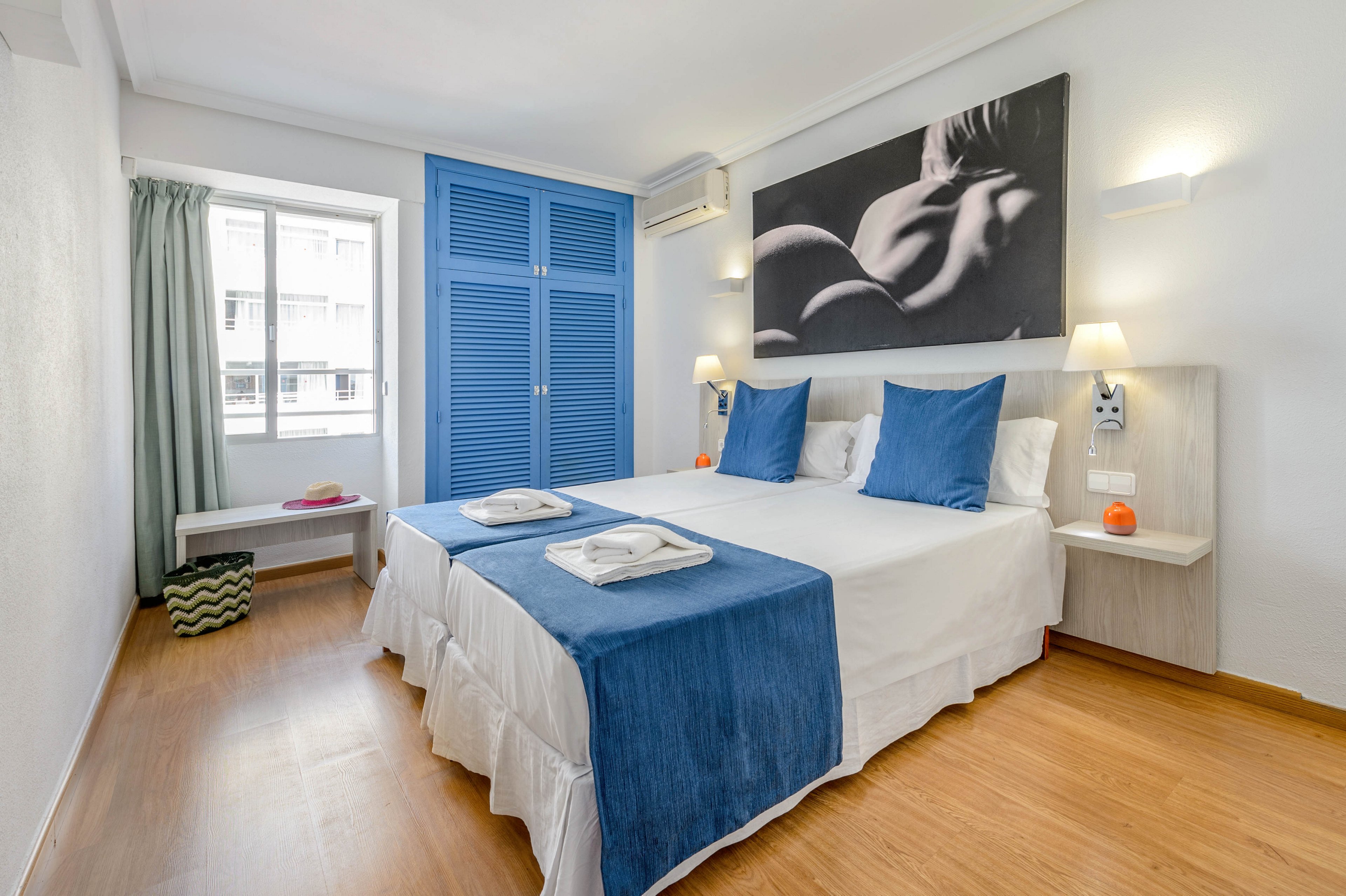 A contemporary hotel room features two single beds joined together, each with crisp white linens and blue throws. The space is well-lit by natural light from a large window, and decorated with blue pillows and a striking monochrome art piece above the bed. The room includes a blue wardrobe, bedside tables with lamps, and minimalistic decor for a clean, inviting atmosphere. No visible text or numbers are present in the image.