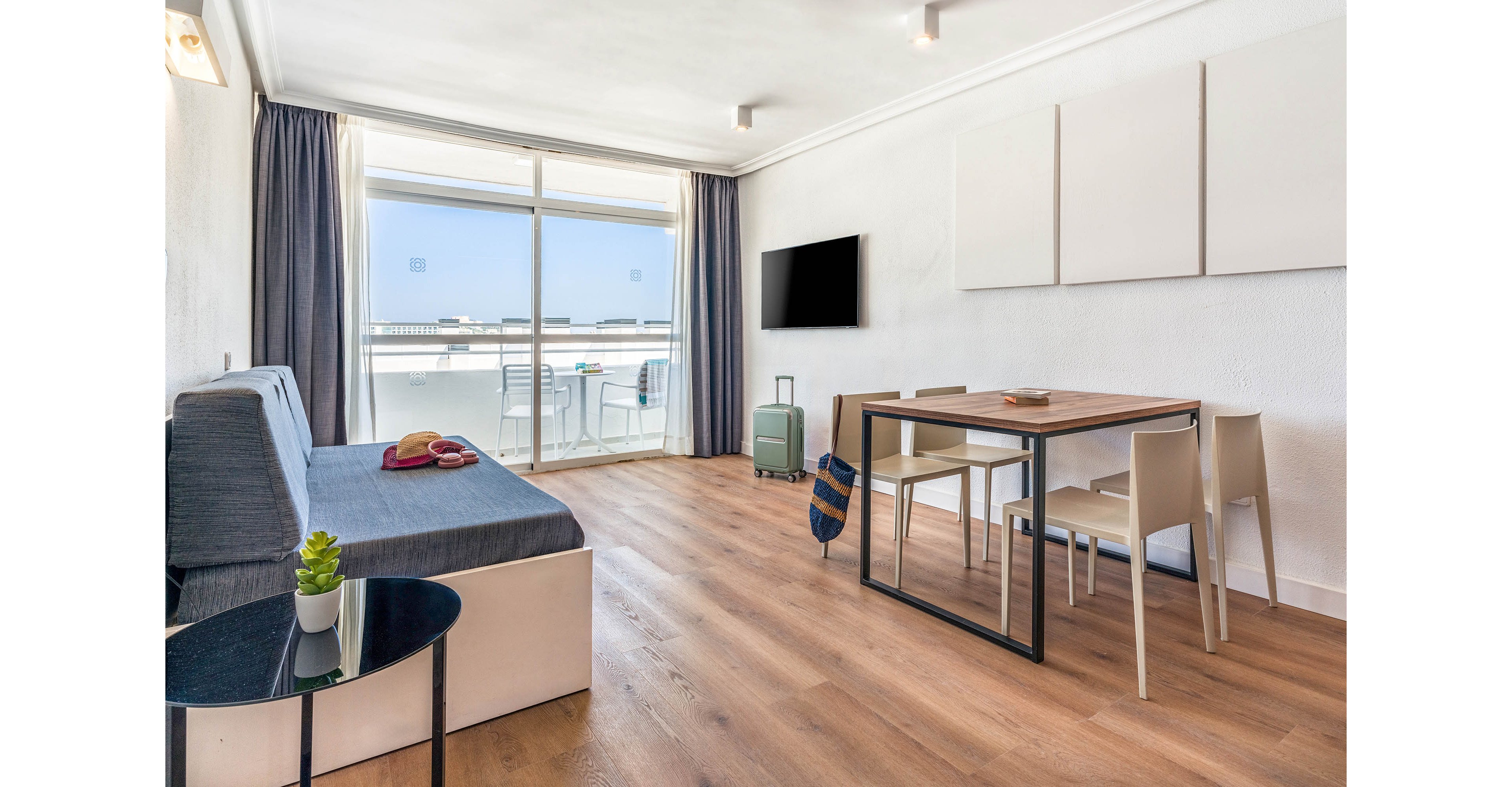 A contemporary apartment living room features a sleek sofa, dining table, and wall-mounted television. Large glass doors open to a sunlit balcony with outdoor seating, creating an airy and inviting atmosphere. The space is decorated in neutral tones with wood flooring and minimalistic decor, including a small potted plant and travel luggage. No visible text or numbers are present in the image.