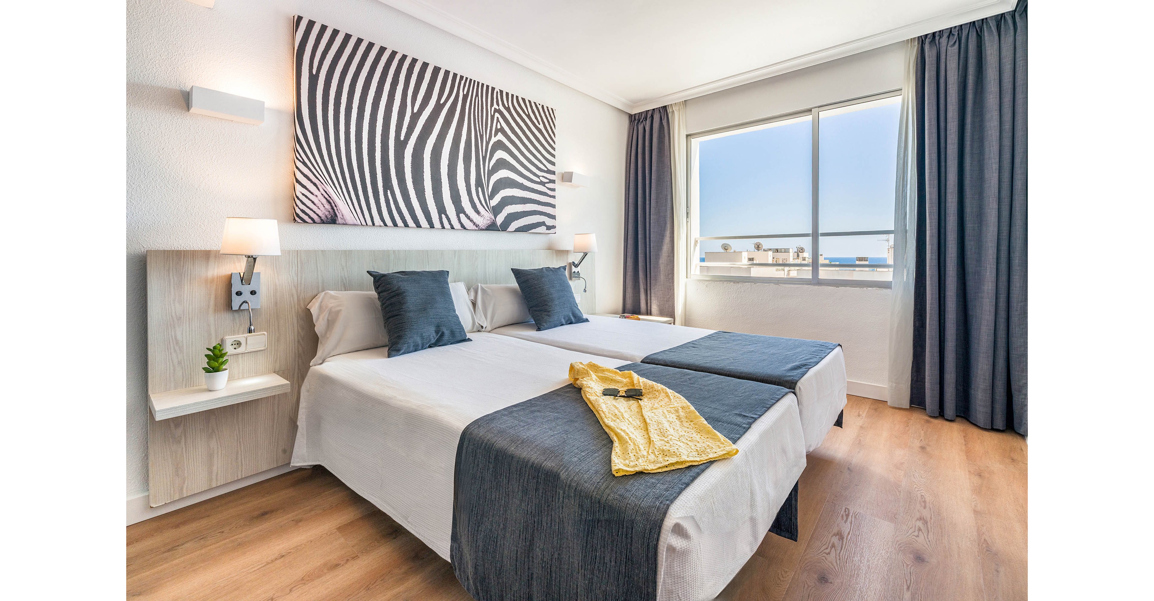 A contemporary hotel room features two neatly made twin beds with gray and white bedding, accented by a yellow throw. The space is decorated with a large zebra-patterned artwork above the headboard and illuminated by wall-mounted lamps. A large window lets in natural light and offers a view of white buildings and blue sky. The room has a clean, minimalist style with wooden flooring and dark curtains. No visible numbers or text are present in the image.