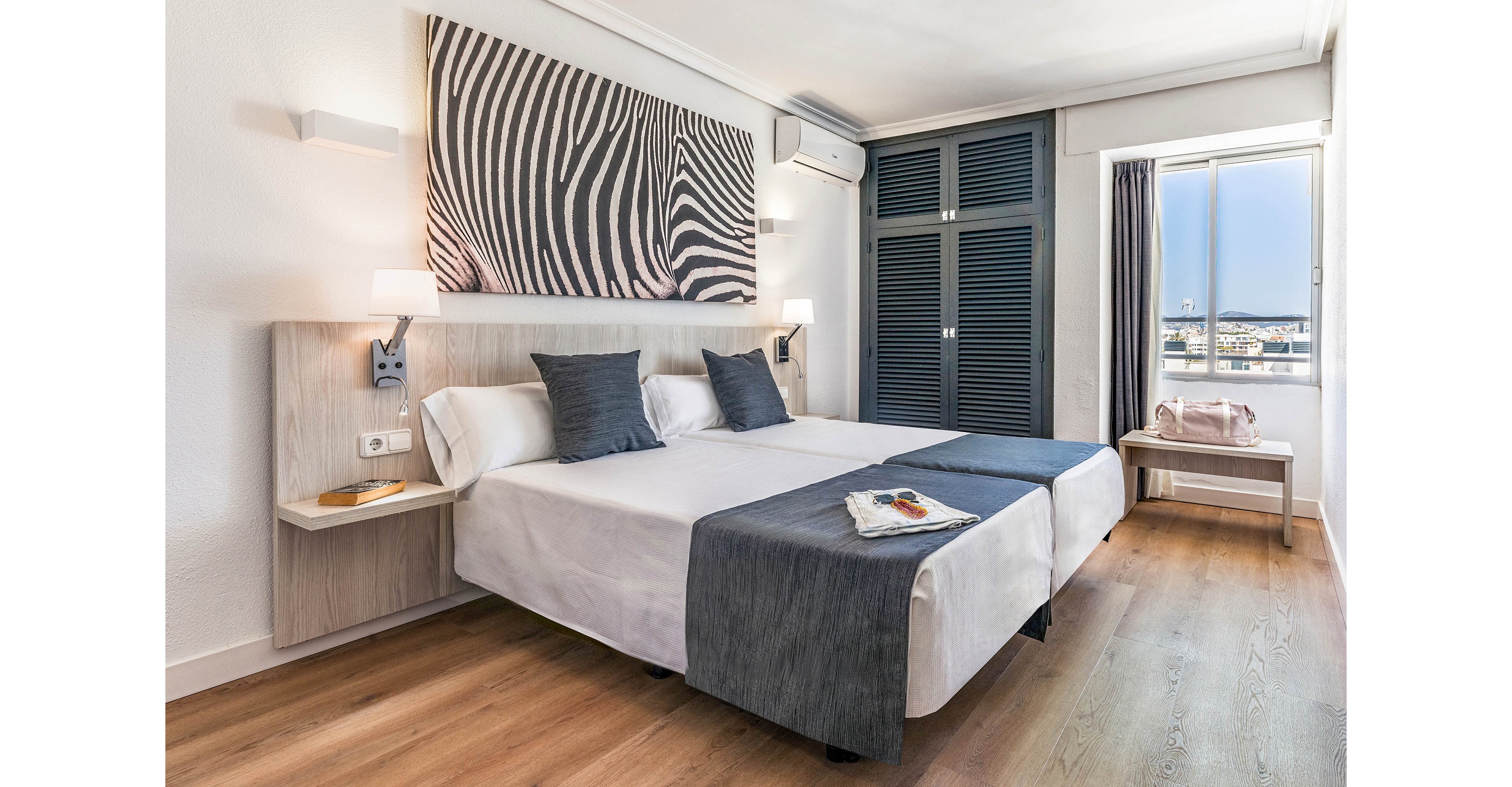 A contemporary hotel room features two twin beds with dark gray runners and pillows, set against a white wall adorned with a large zebra print artwork. The space is bright, with natural light streaming through a window that offers a cityscape view. The room includes a wooden floor, bedside tables, and a wardrobe with black doors. A pink bag sits on a side table, and a magazine is placed on one of the beds. The overall mood is clean, modern, and inviting.
