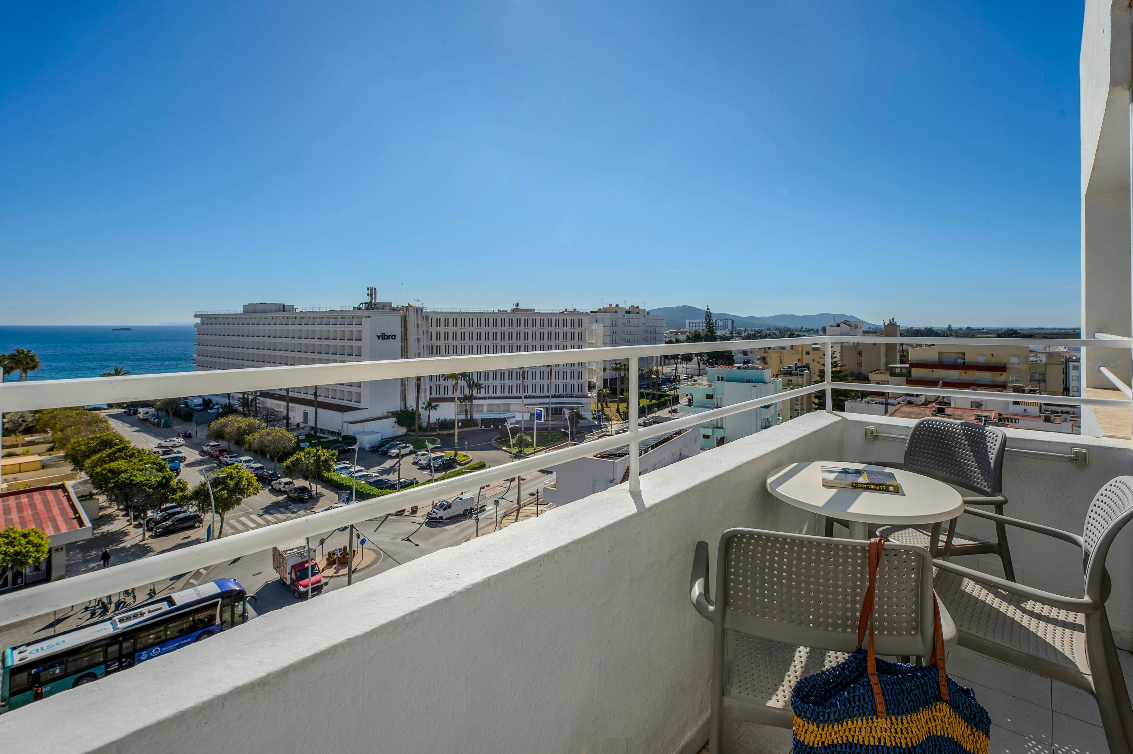 A modern hotel balcony overlooks a vibrant cityscape and the ocean under a clear blue sky. The balcony features a small round table with two chairs, one holding a woven beach bag. Sunlight casts sharp shadows, highlighting the relaxed, vacation atmosphere and the distant mountains on the horizon.