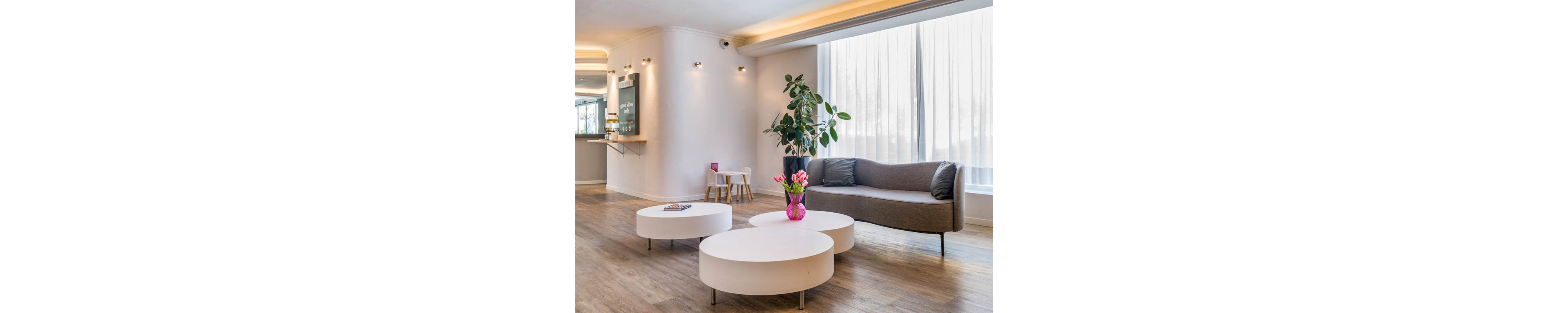 A contemporary lounge area features a sleek gray curved sofa beside large sheer windows. Three low, round white coffee tables sit in the center, one holding a pink vase with fresh tulips and a magazine. A tall potted plant adds greenery to the minimalist interior, while warm recessed lighting highlights the clean white walls. In the background, a small children's table and chairs and a service counter create a welcoming, multi-purpose space.