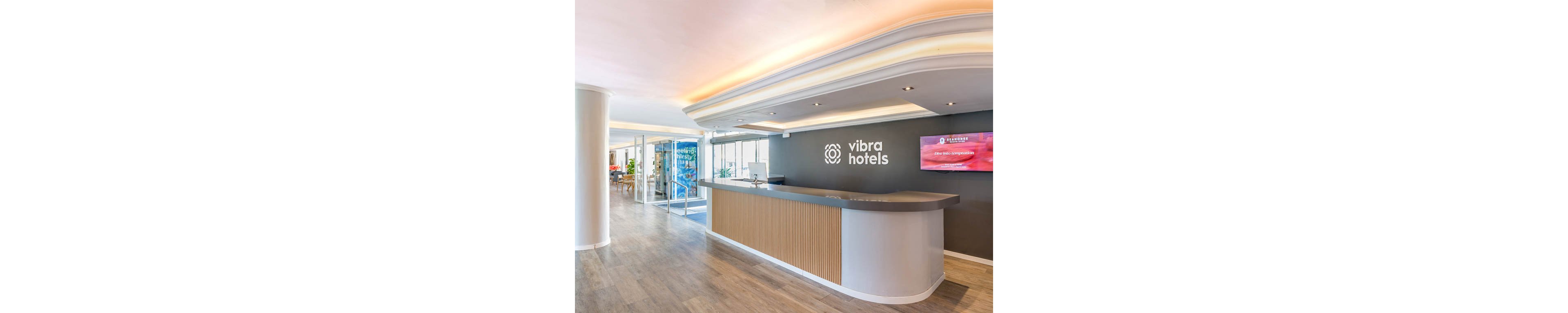 A contemporary hotel reception area features a sleek, curved front desk with wood and neutral finishes. The space is bright and open, with large glass doors leading to an adjacent lounge or lobby area. A wall behind the desk displays the text "vibra hotels" alongside a digital screen showing colorful content. Recessed lighting and a clean ceiling design contribute to the modern, welcoming atmosphere.