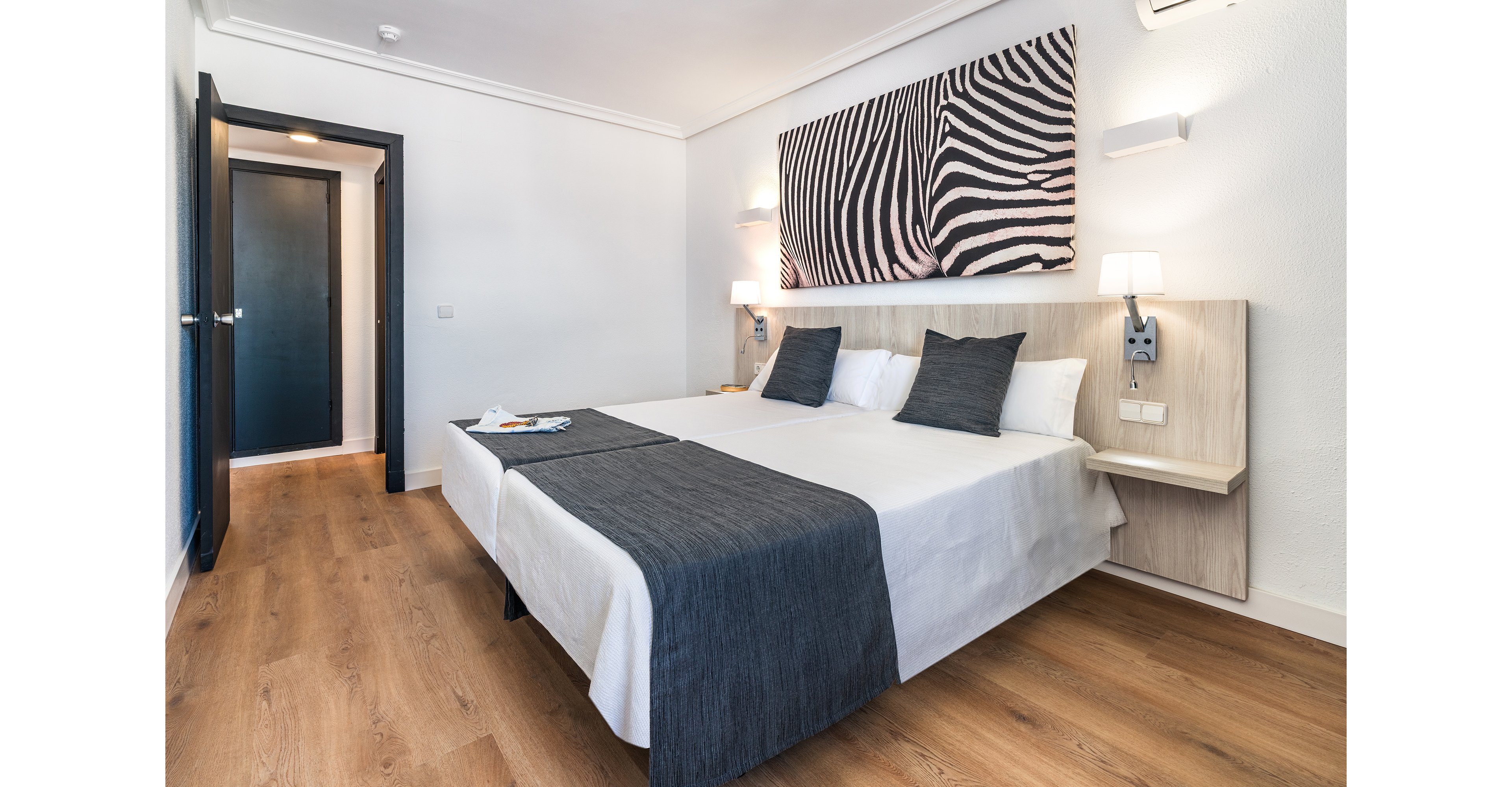 A bright, modern hotel room features two twin beds pushed together with white linens and dark gray throws. The space has light wood flooring, a minimalist bedside table, and wall-mounted lamps on either side of the bed. A large zebra-pattern artwork hangs above the headboard, adding a bold graphic accent. An open doorway reveals a hallway and a dark-colored door, enhancing the sense of depth in the room.