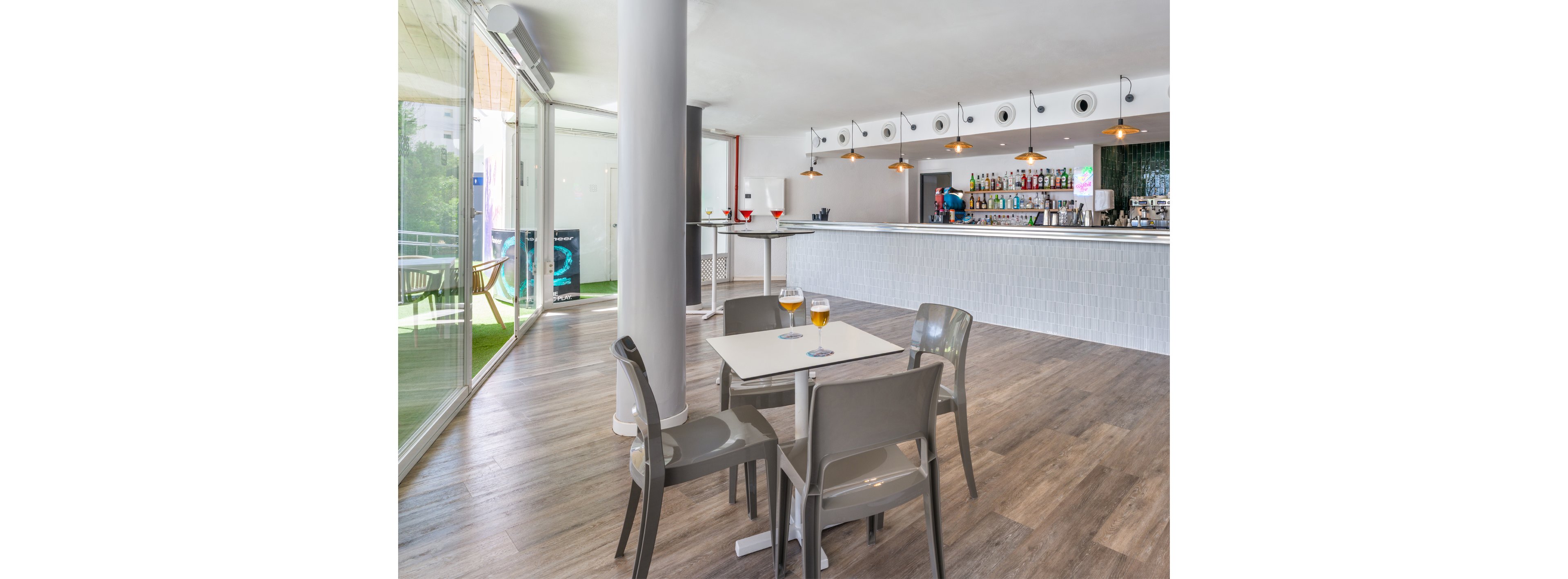 A contemporary indoor bar area features a small square table with two drinks placed on top. The space includes a long counter lined with bottles and glassware, illuminated by hanging pendant lights. Large floor-to-ceiling windows bring in natural light and reveal an outdoor seating area. The clean design, neutral tones, and wooden flooring create a relaxed, modern atmosphere.
