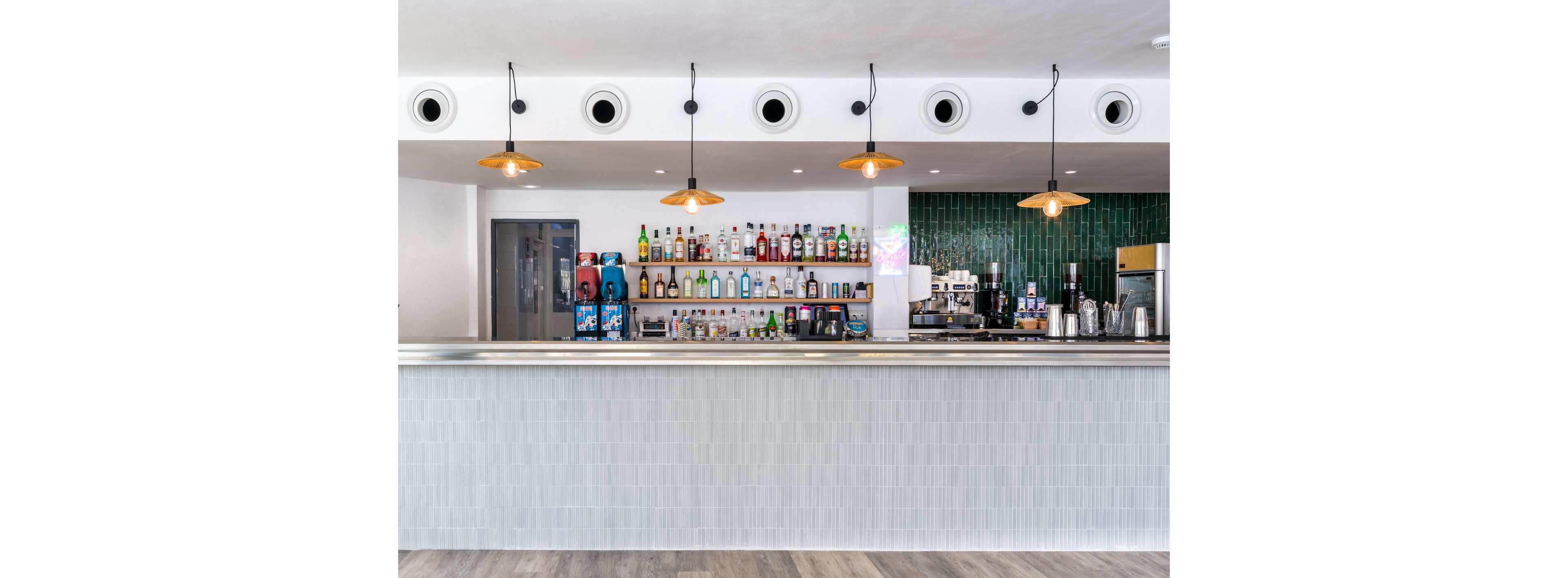 A contemporary bar counter stands in front of a neatly arranged liquor display. Bottles of spirits and mixers line wall-mounted shelves behind the bar, alongside a coffee machine and other bar equipment. Pendant lights hang from the ceiling, casting warm light over the sleek, minimalist interior. The space features clean white walls, green tiled accents, and a light wood floor, creating a bright and modern atmosphere.