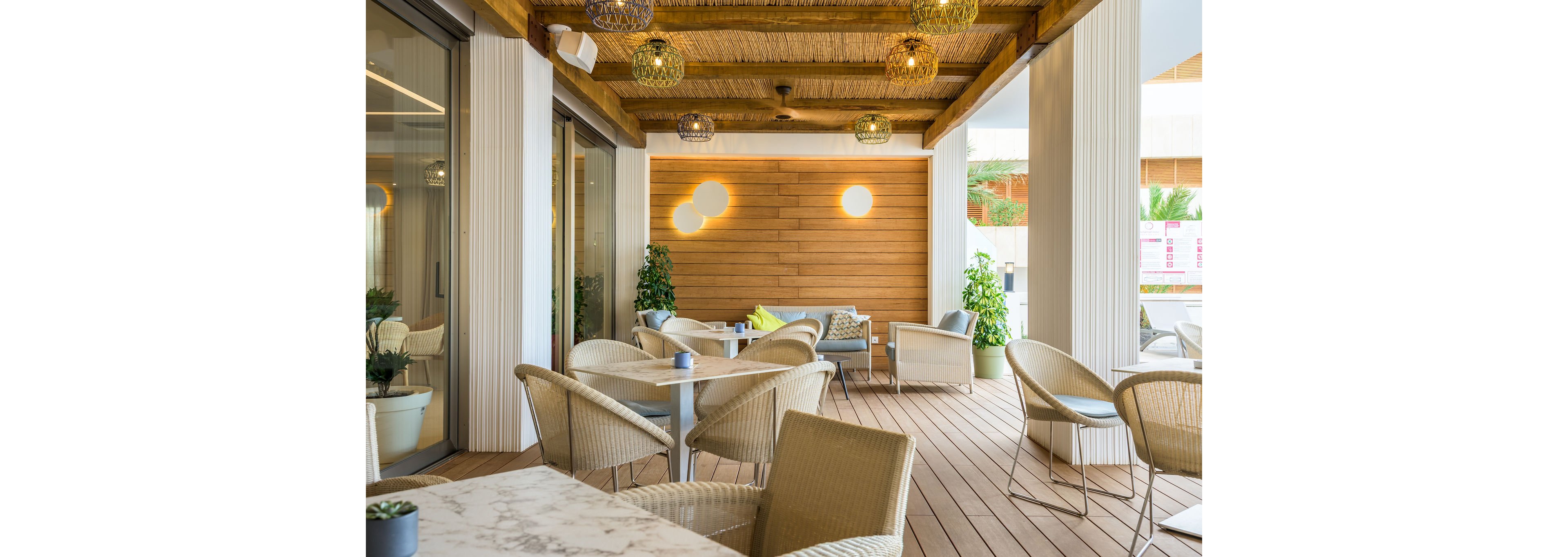 Contemporary outdoor cafe terrace featuring light wicker chairs and round tables on a wooden deck. The space is covered by a wooden pergola with woven ceiling details and decorative hanging lights. A wooden accent wall with circular light fixtures adds warmth, while potted plants bring greenery to the setting. The overall atmosphere is bright, relaxed, and inviting for casual dining or coffee.