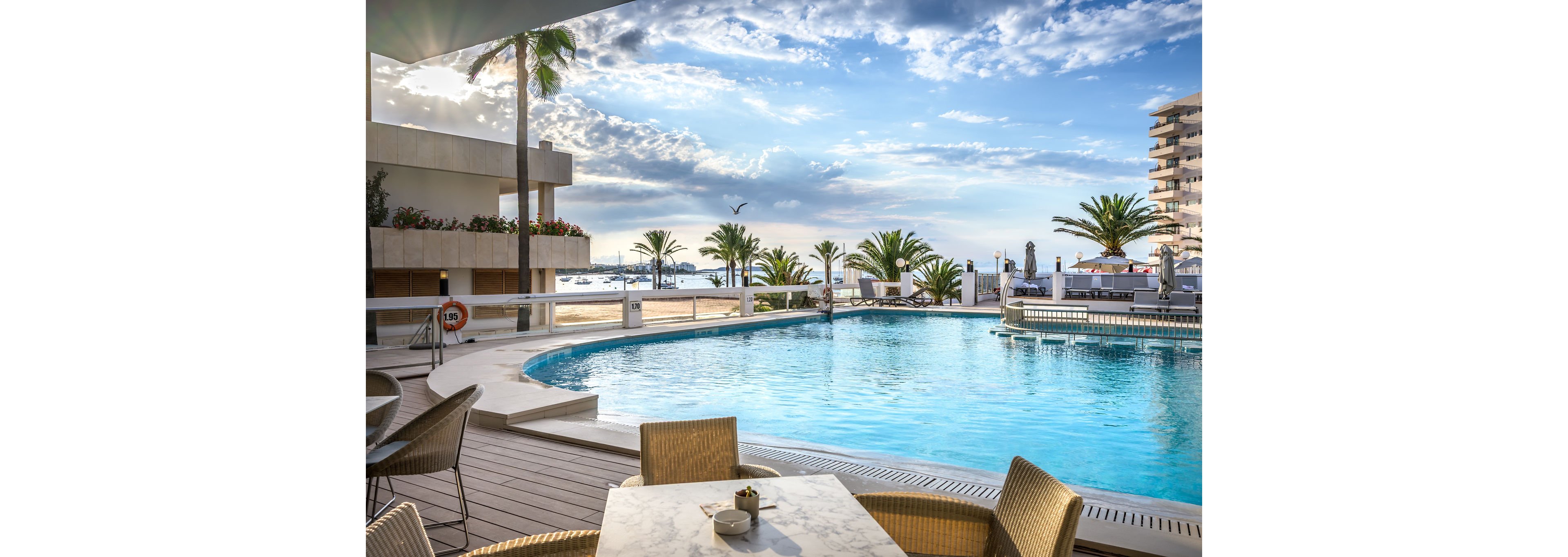 An outdoor swimming pool at a seaside resort is surrounded by a wooden deck and wicker chairs near a marble-topped table. Palm trees and modern buildings frame the view, with the ocean and boats visible in the background. The sky is partly cloudy with warm evening light reflecting on the water, creating a tranquil vacation atmosphere. A lifebuoy and safety sign are visible along the pool edge, emphasizing the leisure yet secure setting.
