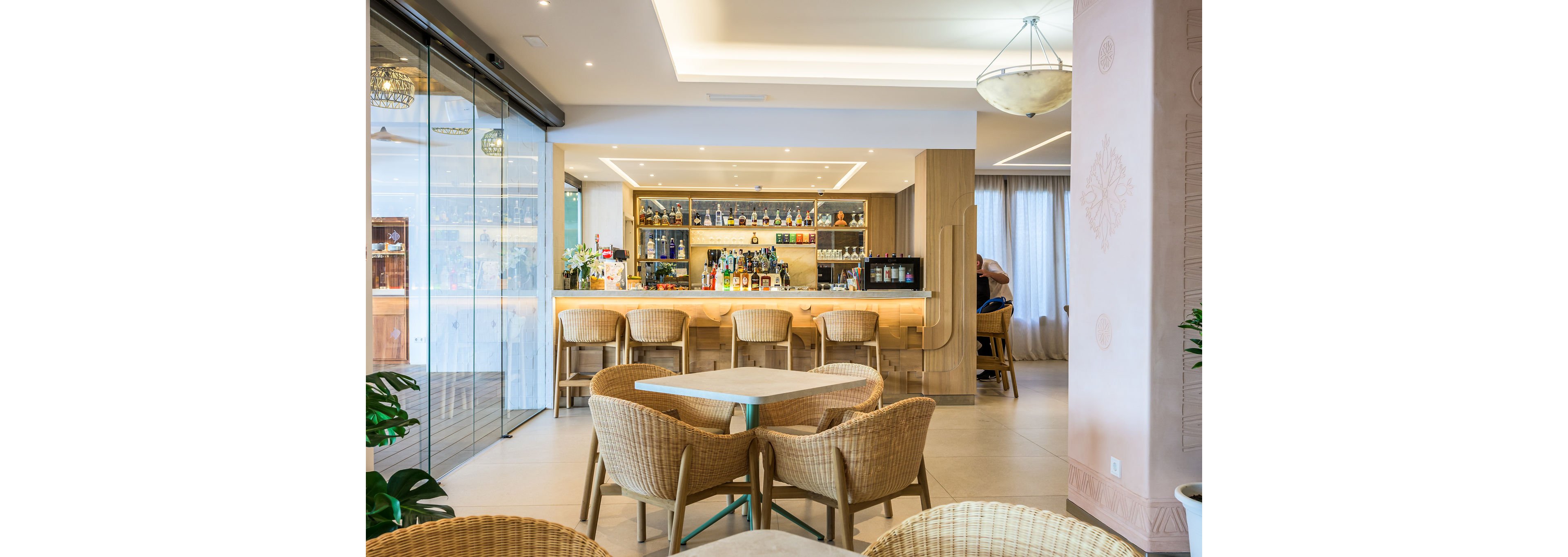 Bright contemporary bar interior featuring a long counter with bottles and glassware neatly arranged on backlit shelves. Wicker chairs surround small tables in the foreground, creating a relaxed lounge area. Large windows and glass doors allow natural light to fill the space, highlighting neutral tones and clean lines. A few people sit at the bar, adding a casual social atmosphere to the modern setting.