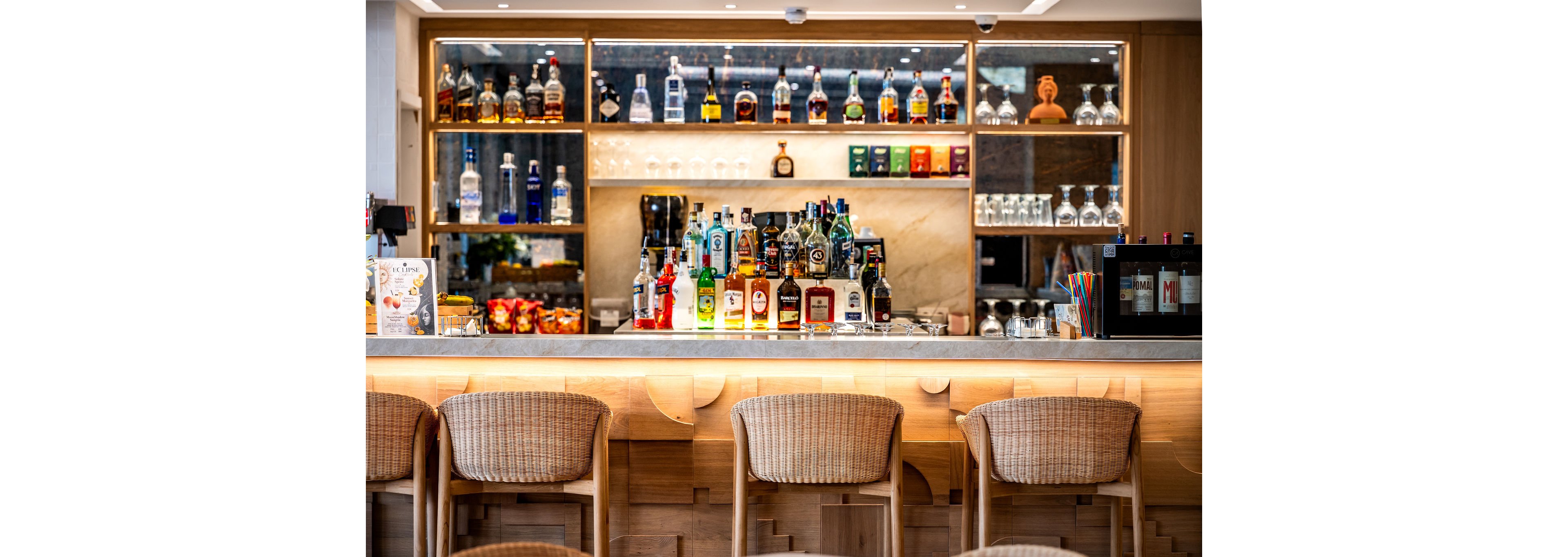 A modern bar counter is lined with four woven stools facing a well-lit liquor display. Numerous bottles of spirits are arranged on shelves and the countertop, with glassware neatly stored to the side. The setting features warm wood tones, accent lighting, and a polished, inviting atmosphere. Colorful straws and small snacks add casual details to the sophisticated interior.