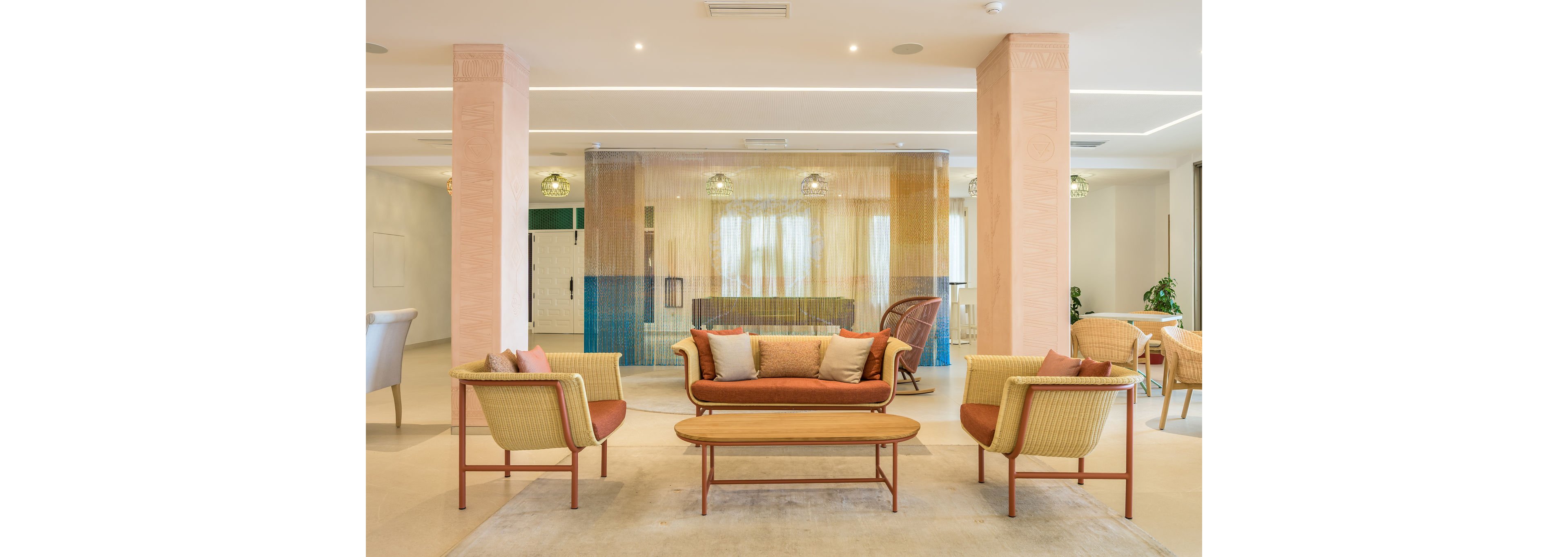 A modern lounge area features a central sofa and woven armchairs arranged around a slim wooden coffee table. The space is open and bright, with pale walls, tall columns, and recessed ceiling lighting. A colorful translucent curtain divides the room, adding soft blue, yellow, and orange tones. Neutral flooring and minimal decor create a calm, welcoming atmosphere suitable for hospitality or residential interiors.