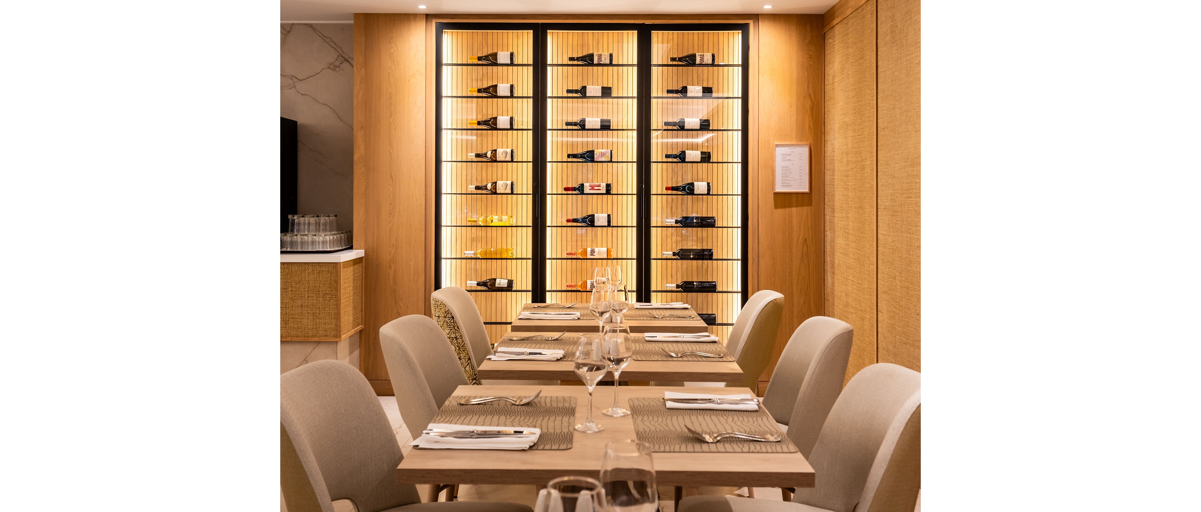 A refined restaurant dining area features a long wooden table set with placemats, napkins, cutlery, and empty wine glasses. Soft, upholstered chairs surround the table, creating a comfortable and inviting atmosphere. In the background, a backlit wall-mounted wine rack showcases multiple bottles arranged neatly on shelves. Warm lighting and neutral tones contribute to a sophisticated, modern interior design. The overall scene suggests a high-end venue prepared for an intimate meal or special occasion.