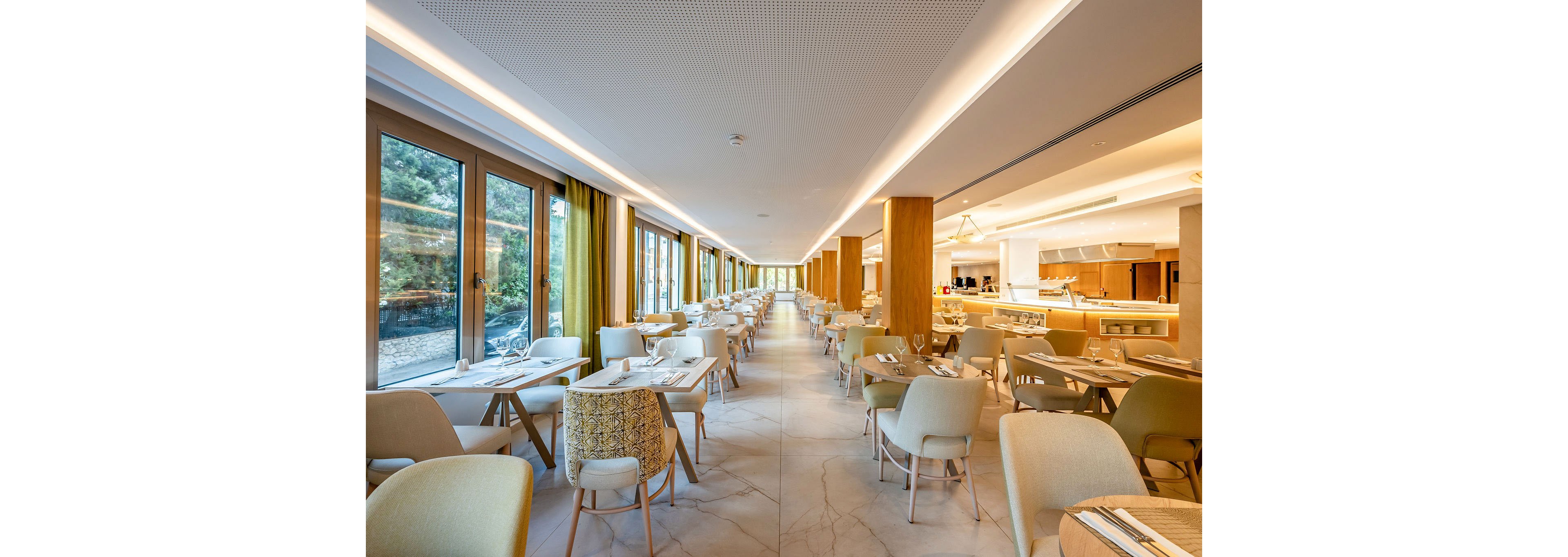 Spacious contemporary restaurant interior with rows of neatly arranged tables and chairs. Large floor-to-ceiling windows line one side, allowing natural light to fill the space and reveal greenery outside. Warm wood accents, soft neutral upholstery, and yellow curtains create a welcoming atmosphere. The open-plan layout leads toward a bright service or buffet area at the far end.