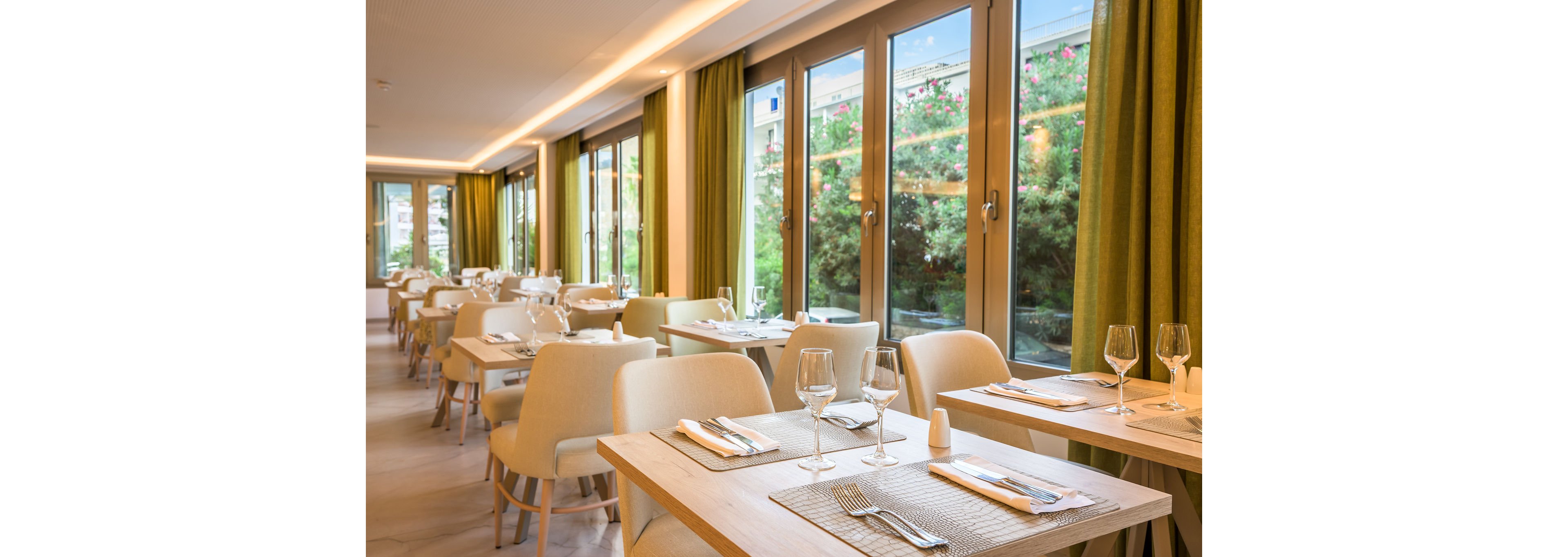 Contemporary restaurant tables are neatly set with glassware, cutlery, and placemats in a bright dining room. Large floor-to-ceiling windows line one side, revealing greenery and flowers outside. Soft upholstered chairs and light wood tables create a clean, minimalist look. Warm lighting and green curtains contribute to a calm, inviting atmosphere for guests.