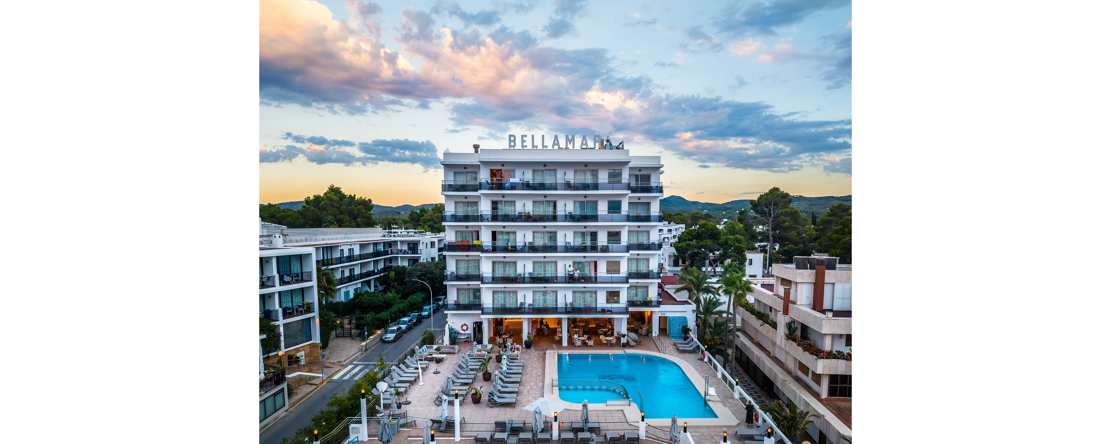 A multi-story Bellamar hotel stands prominently with a large outdoor swimming pool in front. Sun loungers and umbrellas surround the pool area, suggesting a relaxing vacation setting. The building is framed by nearby hotels, trees, and distant hills under a colorful evening sky. Warm lights from the ground floor create an inviting atmosphere for guests.