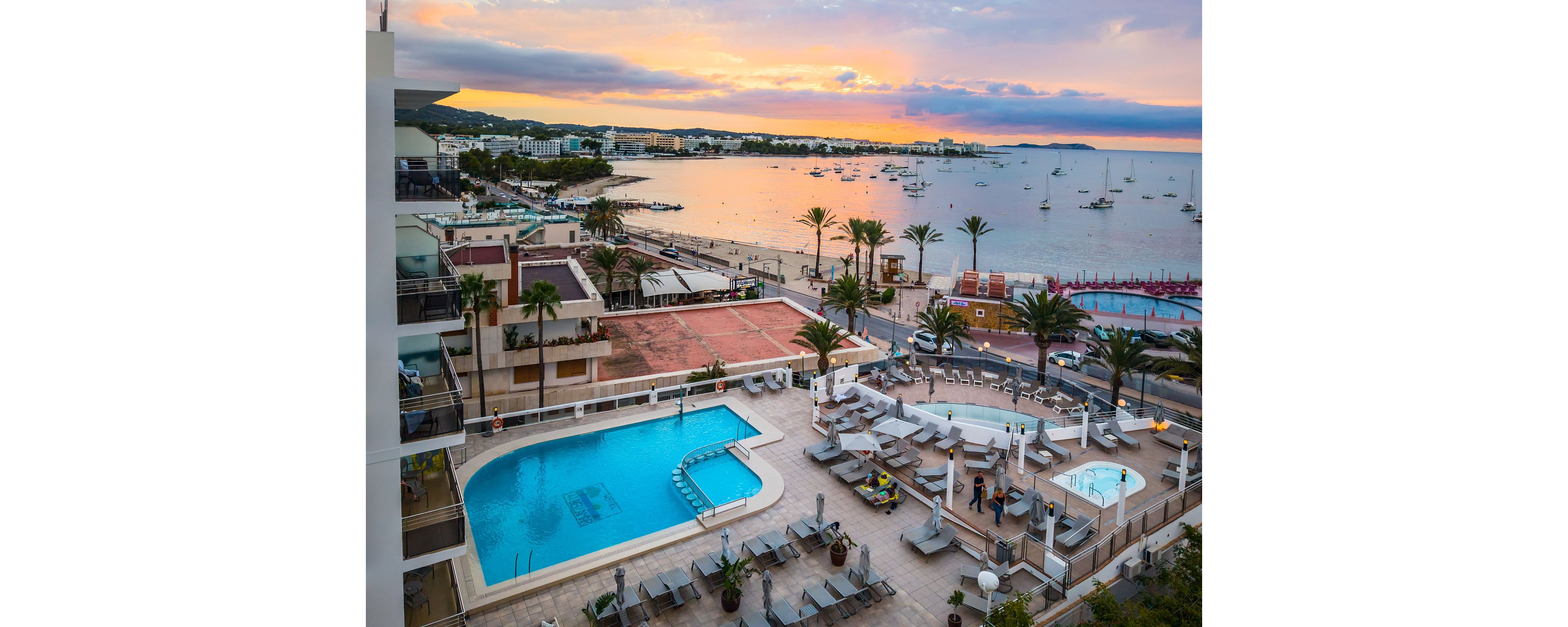 A coastal resort features a large outdoor swimming pool and sun loungers on multiple terraces. Guests relax near the pool and seating areas while overlooking a calm bay dotted with boats. Palm trees line the waterfront promenade and modern buildings frame the shoreline. The sky glows with warm sunset colors, reflecting softly on the water and creating a tranquil vacation atmosphere.