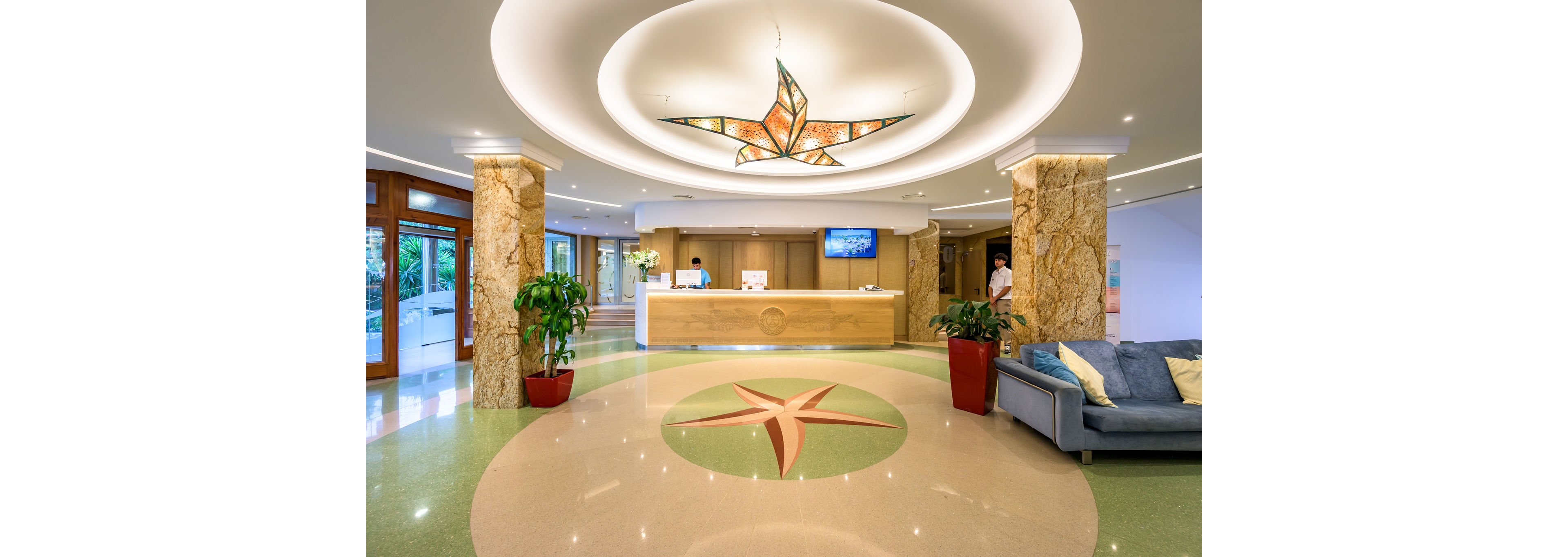 A spacious hotel lobby features a central circular floor design with a star motif leading toward a wooden reception desk. Warm marble columns and recessed ceiling lighting create an inviting atmosphere. A staff member stands behind the counter while another person approaches, and a large digital screen glows behind the desk. Potted plants and a blue sofa with cushions add color and comfort to the contemporary interior.