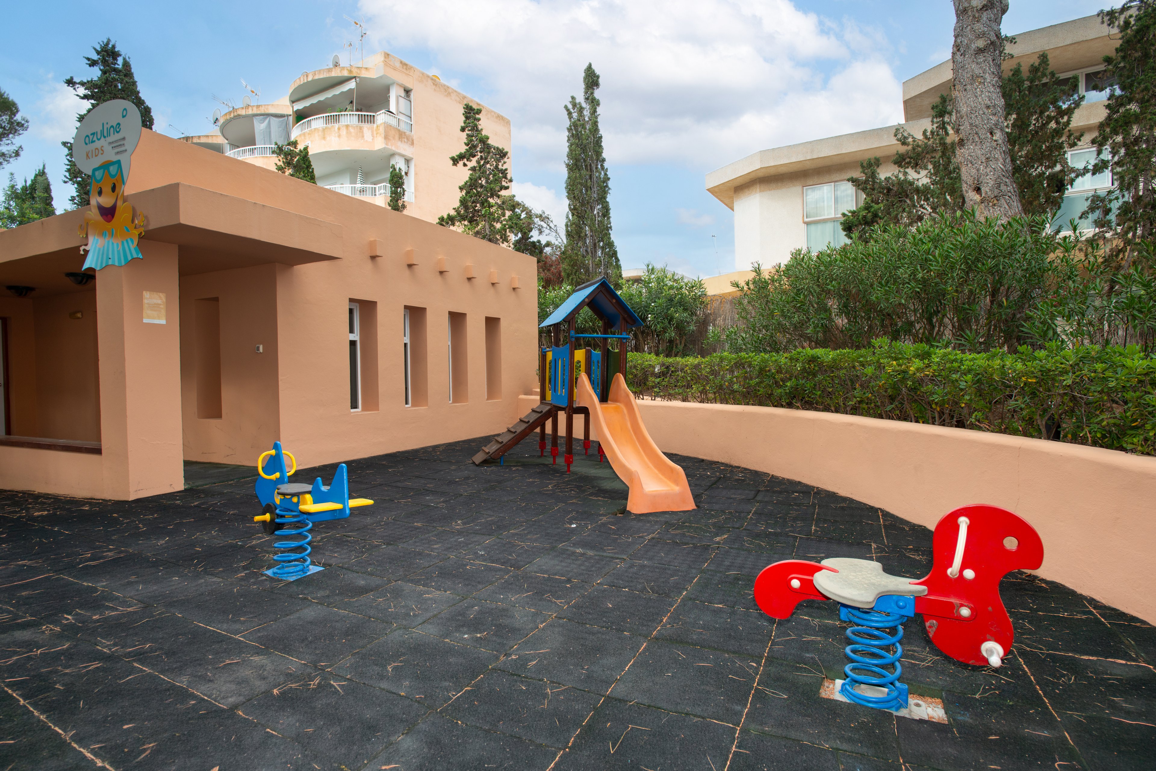 An outdoor playground features colorful play equipment beside a peach-colored building. A small play structure with a slide stands on a rubber safety surface. Two spring riders shaped like animals are positioned in the foreground. Trees, shrubs, and modern buildings surround the play area under a partly cloudy sky.