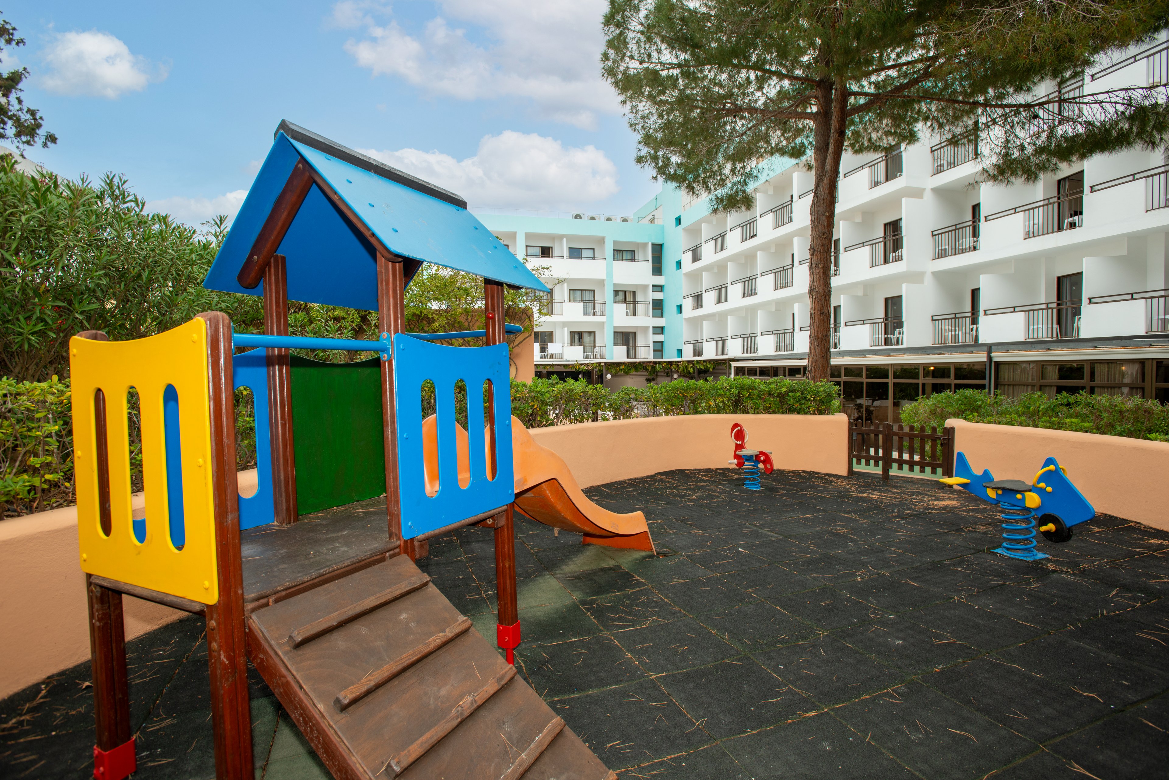 A small playground with a colorful wooden play structure stands in the foreground. The structure includes steps, a slide, and panels painted in bright yellow, blue, green, and red. Nearby are spring-mounted rocking toys set on a dark rubber safety surface. Behind the playground, a multi-story white apartment or hotel building with balconies is visible, surrounded by trees and greenery under a partly cloudy sky.