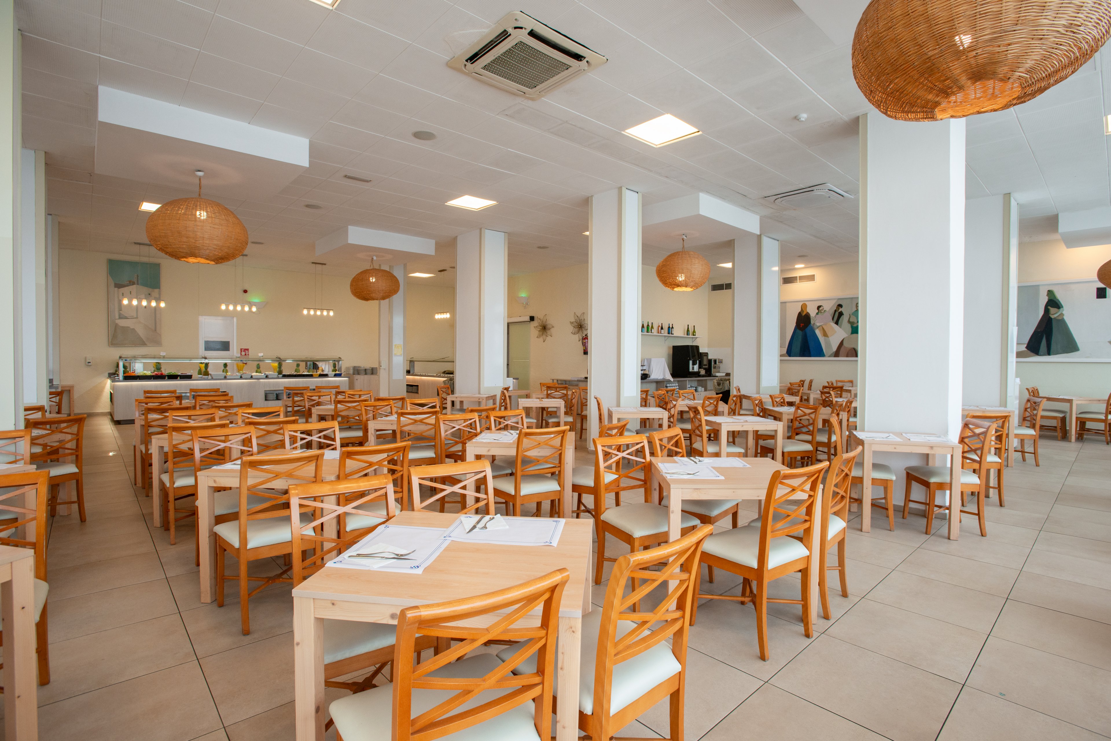 Spacious cafeteria interior featuring rows of light wooden tables and chairs. The room is brightly lit with recessed ceiling lights and large wicker pendant lamps. A long self-service counter is visible at the back with food displays and equipment. White walls, tiled flooring, and minimal decor create a clean, contemporary dining atmosphere.