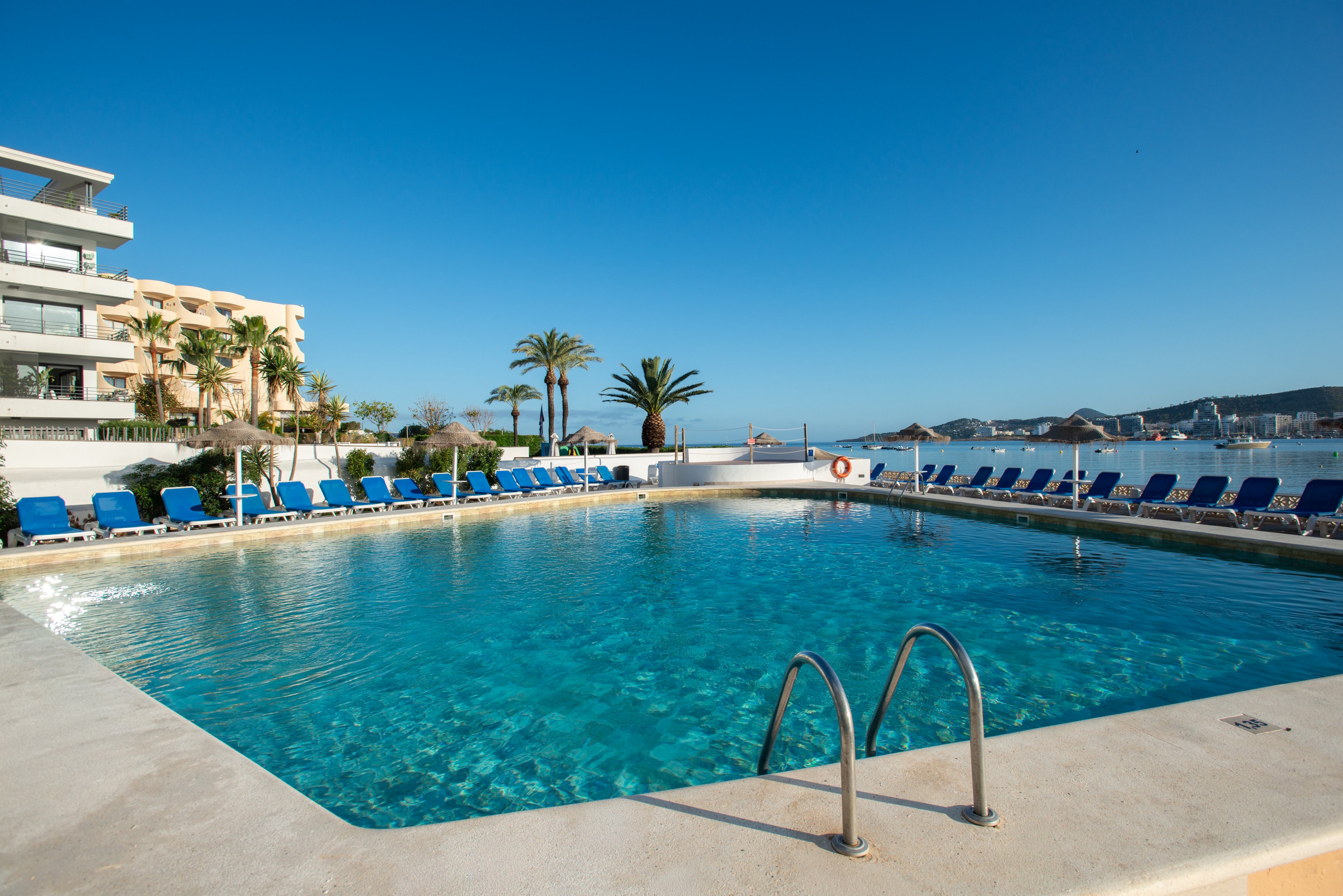 Large outdoor swimming pool at a seaside resort under a clear blue sky. Rows of blue lounge chairs surround the pool, suggesting a relaxing vacation setting. Palm trees and modern hotel buildings frame the scene, with calm water and distant boats visible beyond the pool. The bright sunlight and clear water create a tranquil, summery atmosphere.