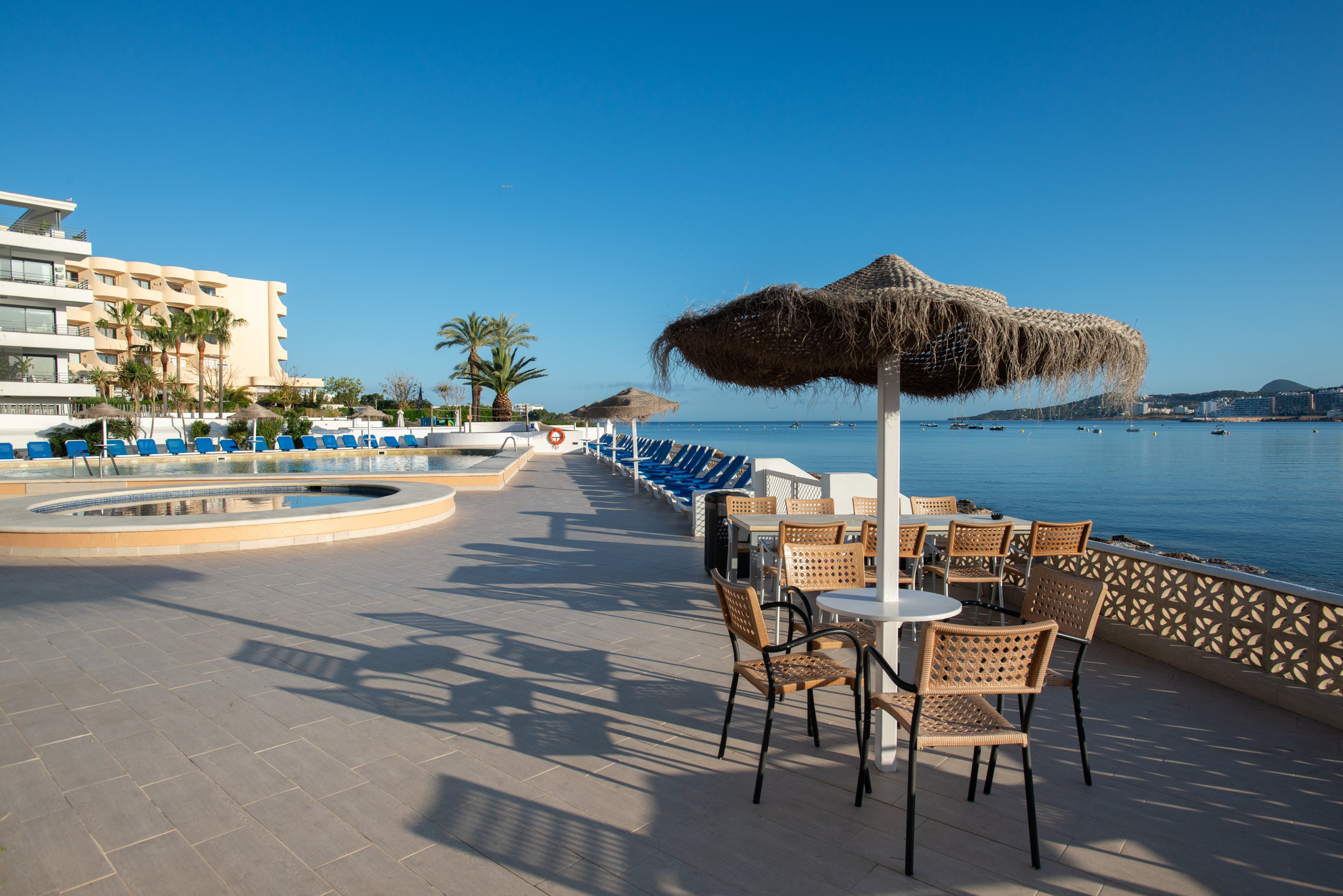 A quiet seaside resort terrace overlooks calm blue water under a clear sky. Empty lounge chairs line the poolside area beside a circular swimming pool. A small table with wicker chairs sits under a thatched umbrella casting long shadows on the pavement. Modern hotel buildings and palm trees frame the waterfront, creating a peaceful vacation atmosphere.