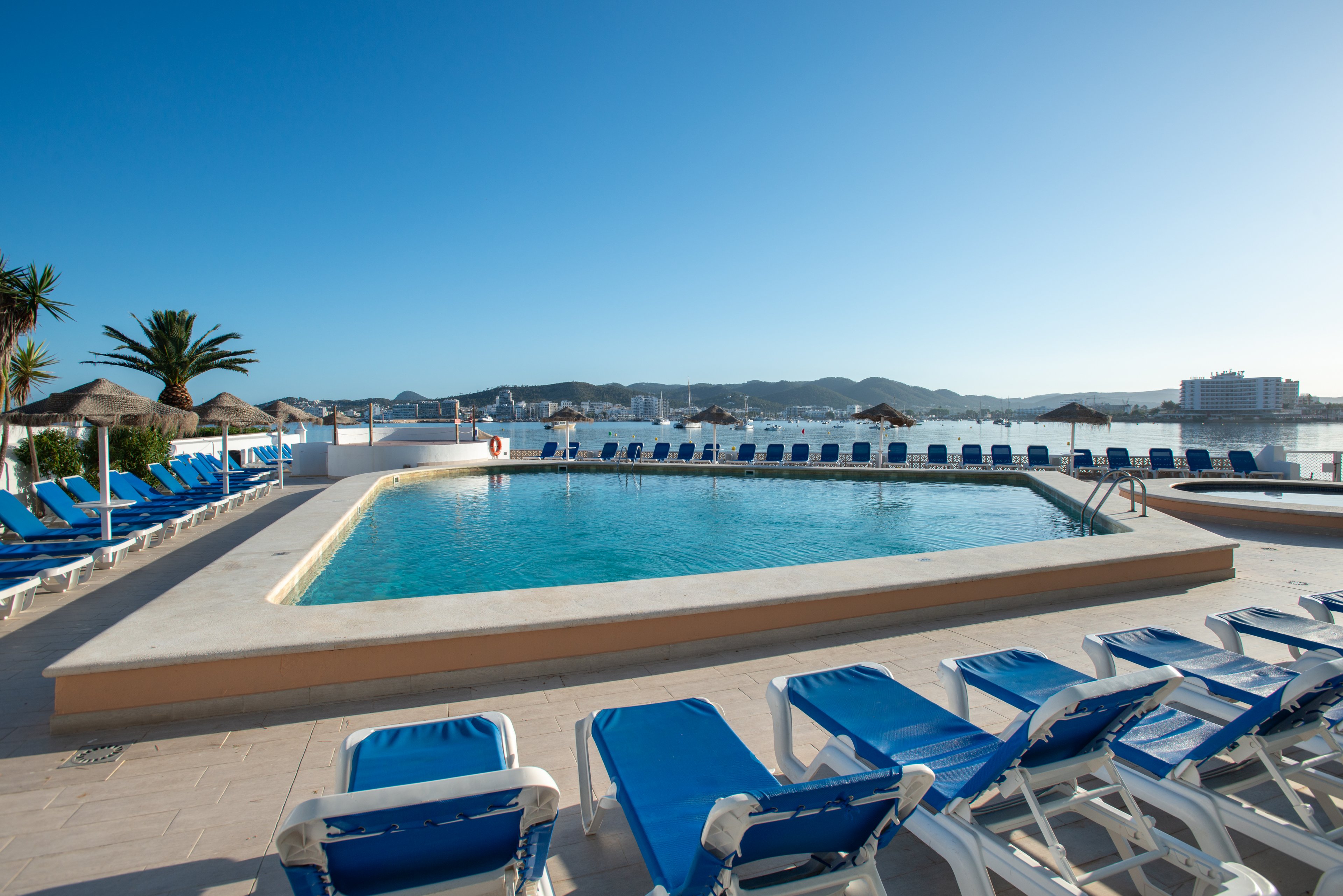 A large outdoor swimming pool sits at the center of a resort terrace, surrounded by rows of blue and white sun loungers. The scene overlooks a calm waterfront with boats and distant buildings under a clear blue sky. Palm trees and thatched umbrellas add a tropical holiday atmosphere. The area appears quiet and empty, suggesting early morning or off-peak relaxation time.