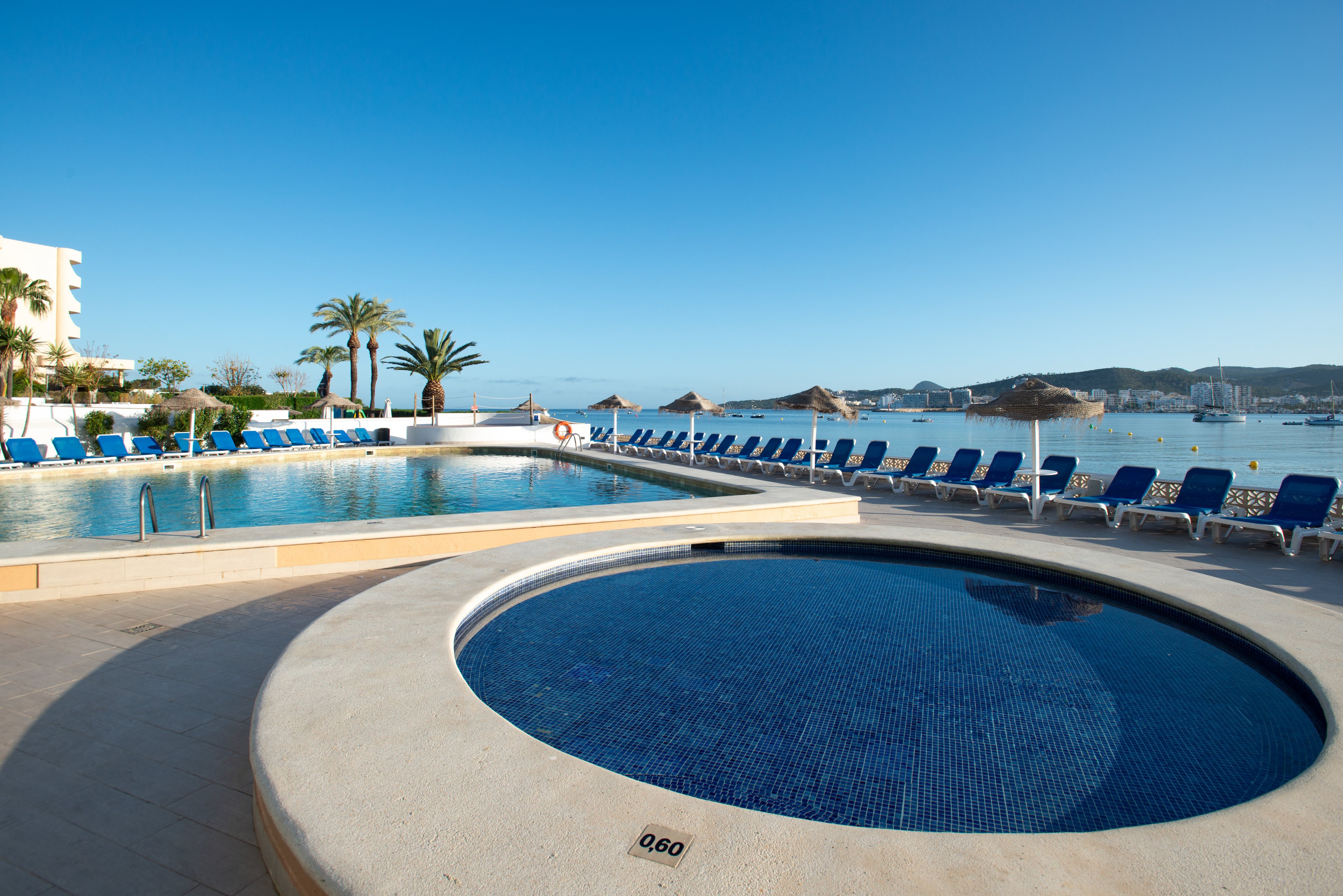 A circular shallow pool in the foreground overlooks a larger rectangular swimming pool at a seaside resort. Rows of blue lounge chairs line the pool deck, facing the calm water and distant shoreline. Palm trees, straw parasols, and clear blue skies create a tranquil vacation atmosphere. The scene is empty of people, emphasizing relaxation and peaceful early morning light.