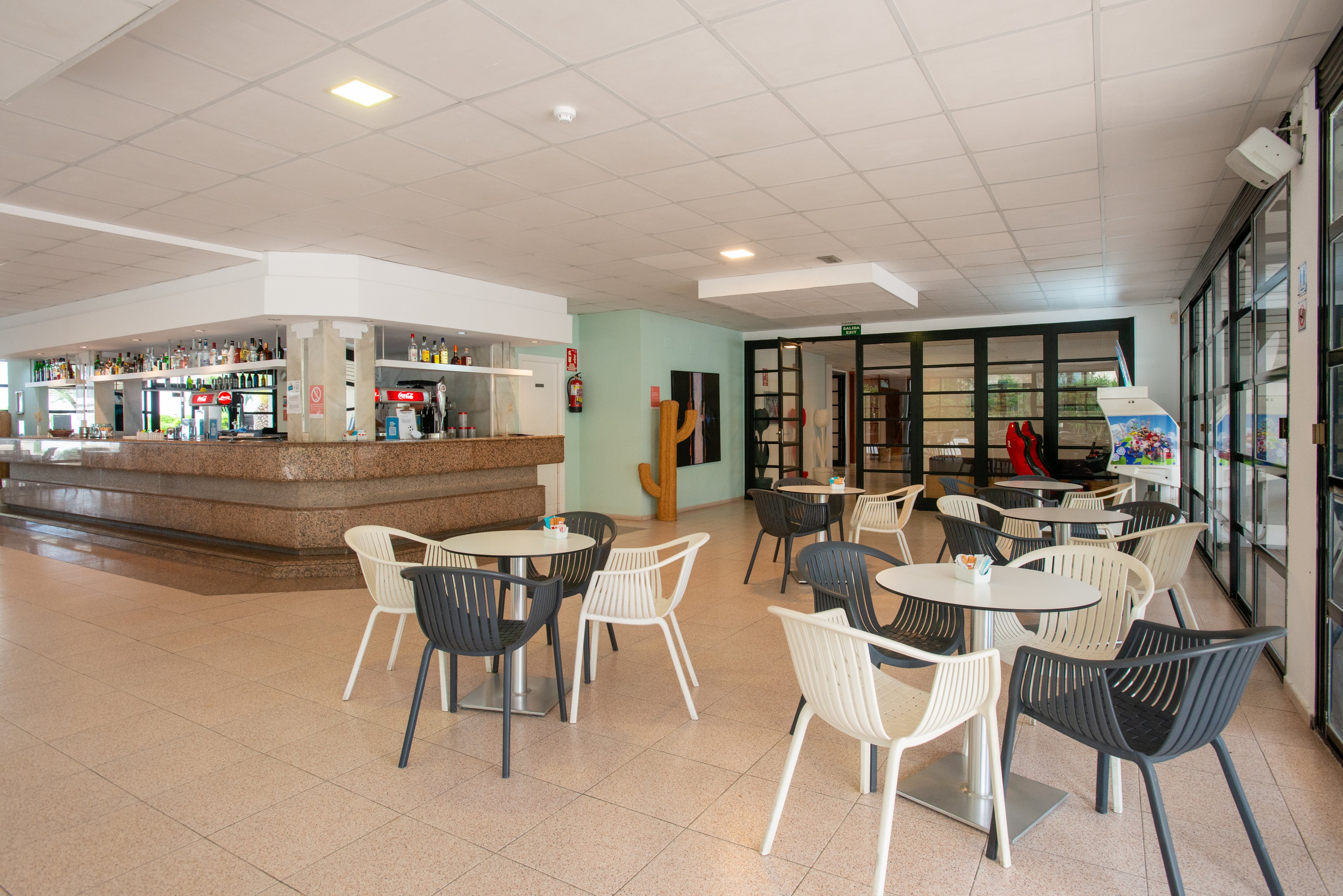 Spacious indoor bar area with several round tables and modern plastic chairs arranged on a tiled floor. The bar counter in the background is stocked with various bottles and drinks, with no customers present. Large windows and glass doors let in natural light, creating an open and airy atmosphere. The overall setting feels clean, casual, and ready for guests in a hotel or resort environment.