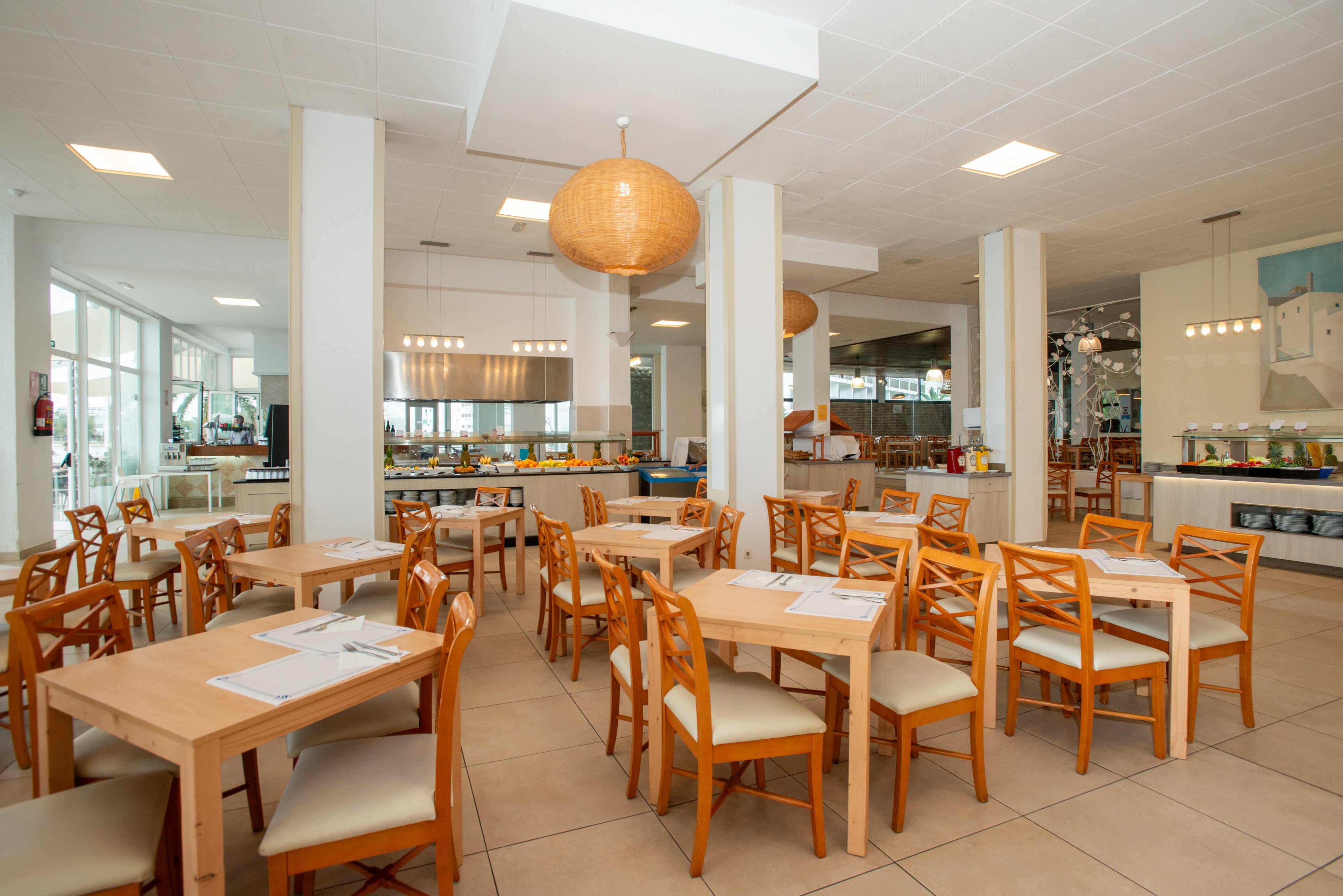 Spacious restaurant dining room with light wooden tables and chairs is shown neatly arranged. The setting features large windows, tiled floors, and white columns creating an airy, open atmosphere. Warm pendant lights hang from the ceiling above the seating area, while a buffet counter and service area are visible in the background. The overall scene appears clean, organized, and ready to welcome guests.