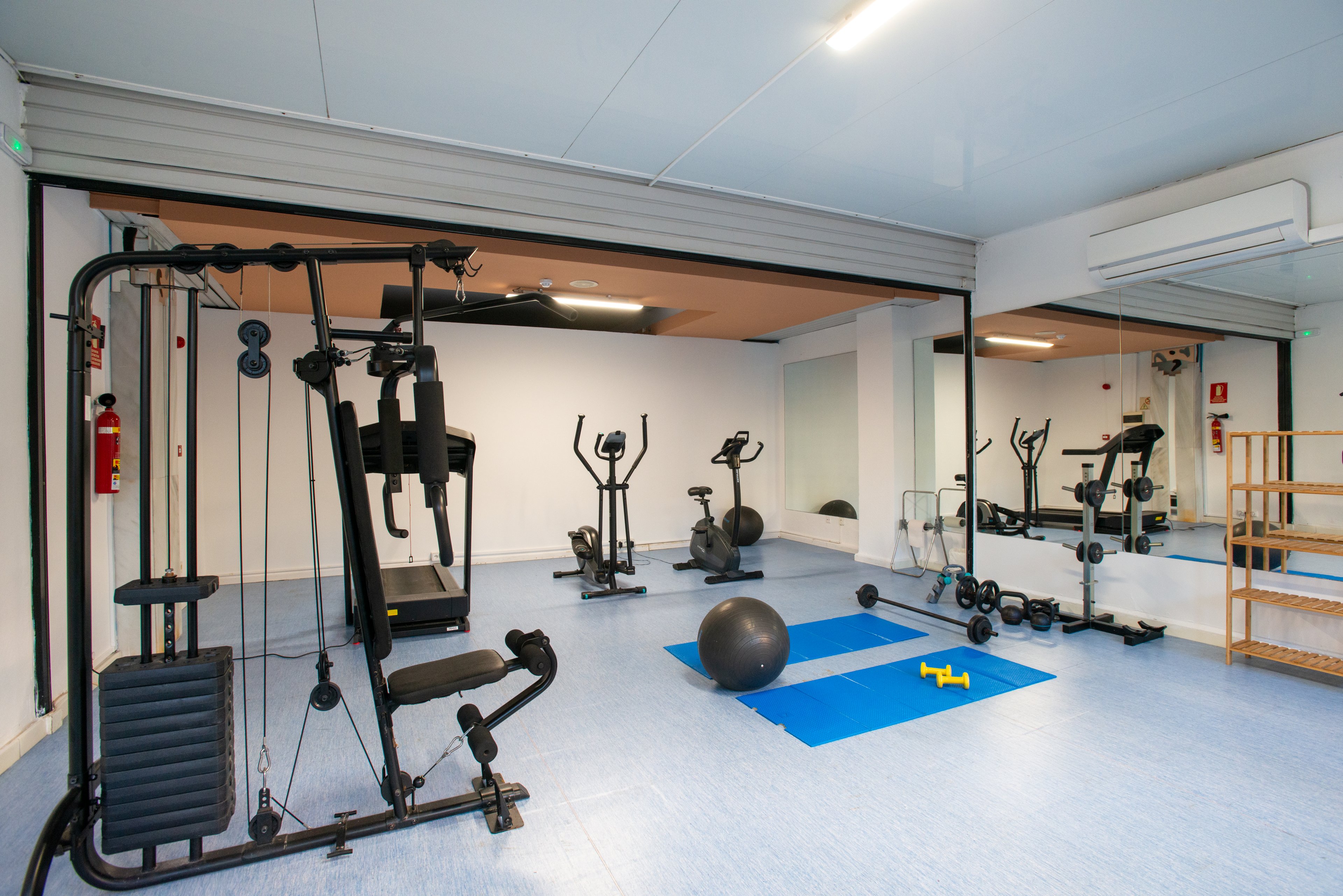 Spacious indoor gym featuring a multi-station weight machine and several cardio trainers. The room includes exercise bikes, an elliptical trainer, and a treadmill arranged in front of large mirrors. On the floor, a blue exercise mat, stability ball, and small dumbbells create a dedicated workout area. The clean, bright space has light blue flooring, white walls, and ceiling-mounted lighting for a fresh, inviting atmosphere.