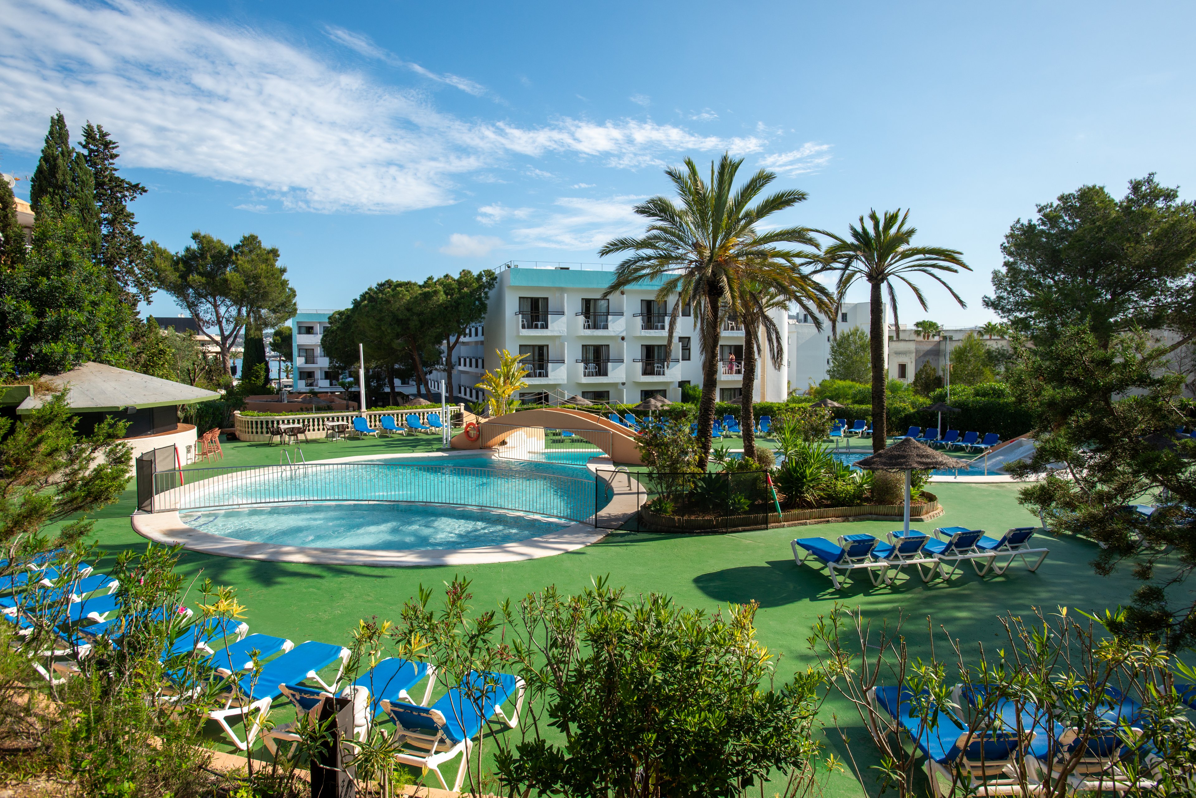 An outdoor resort swimming pool is surrounded by blue sun loungers and lush greenery. A small arched bridge crosses part of the pool, adding interest to the leisure area. White low-rise apartment-style buildings stand in the background under a bright blue sky with scattered clouds. Tall palm trees and other Mediterranean vegetation create a relaxed holiday atmosphere.