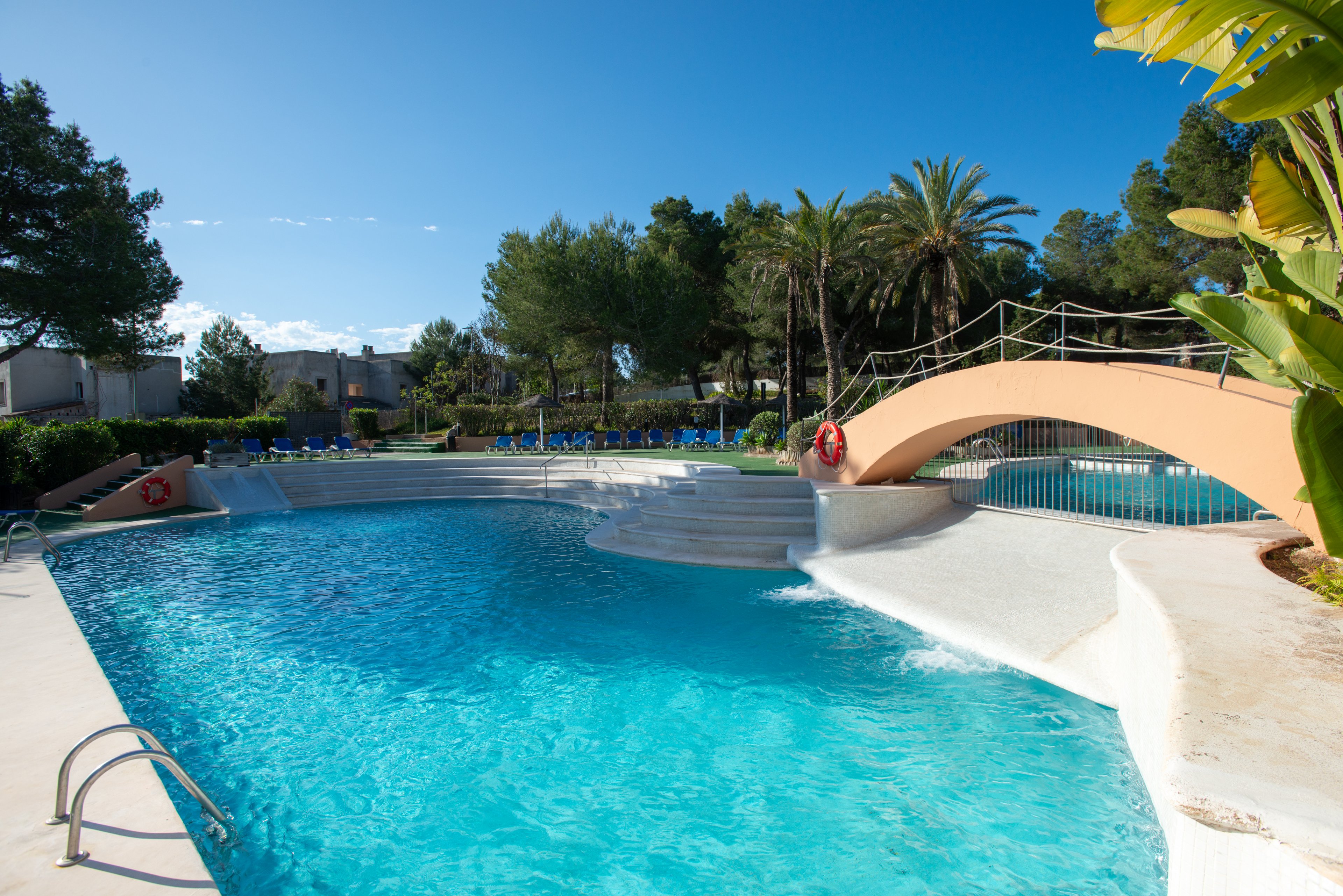 An outdoor resort swimming pool sparkles under a clear blue sky. A small arched bridge crosses part of the pool, with life rings attached to its sides for safety. Sun loungers and umbrellas line the far edge, surrounded by lush green trees and palm trees. The scene suggests a quiet, relaxing holiday atmosphere in a warm climate.