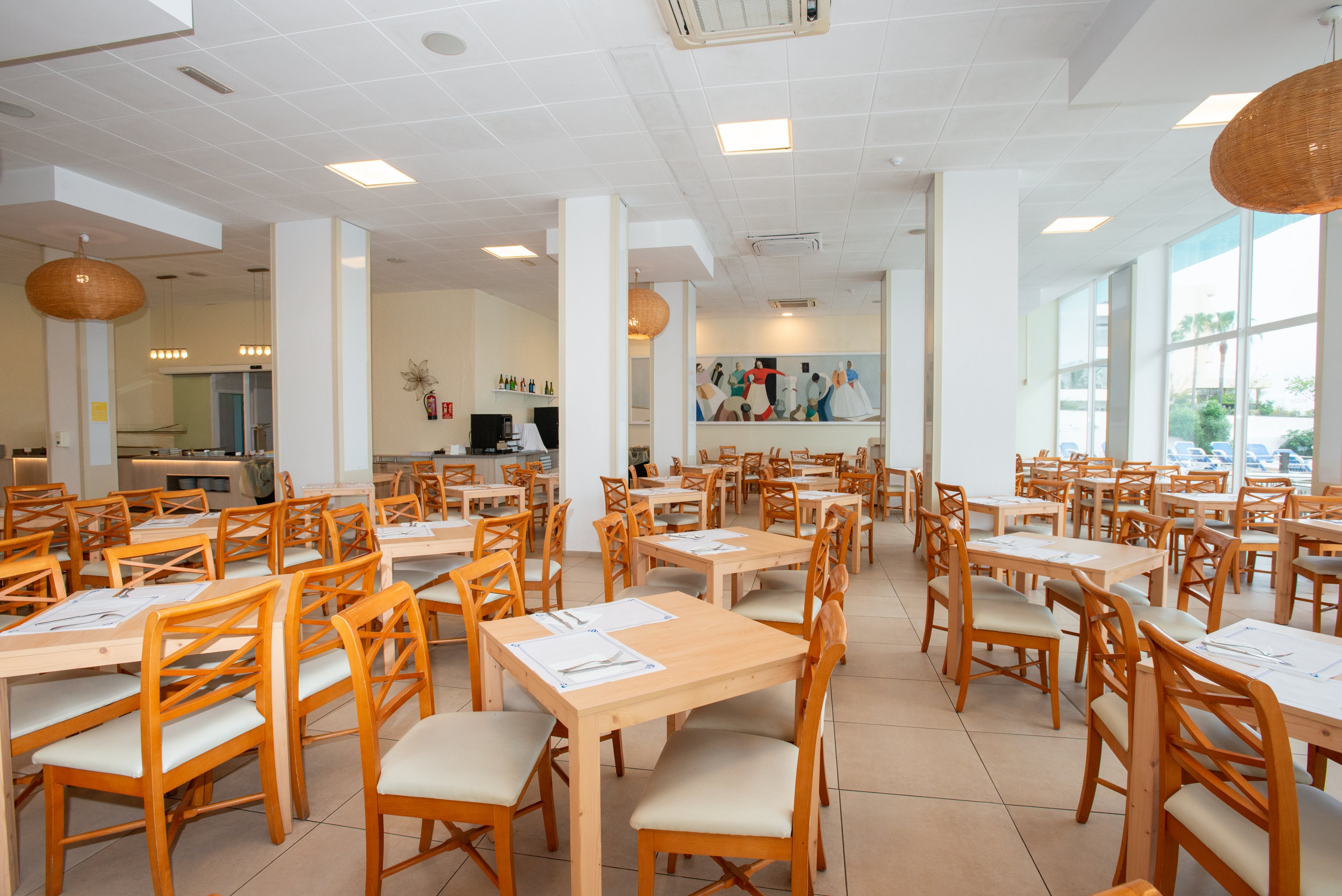 Spacious restaurant dining room with many wooden tables and chairs arranged in neat rows. The setting appears clean and modern, with large windows letting in natural daylight. Neutral walls, tiled floors, and simple ceiling lights create an airy, minimalist atmosphere. A colorful wall painting and woven pendant lamps add subtle decorative accents to the otherwise uncluttered space.
