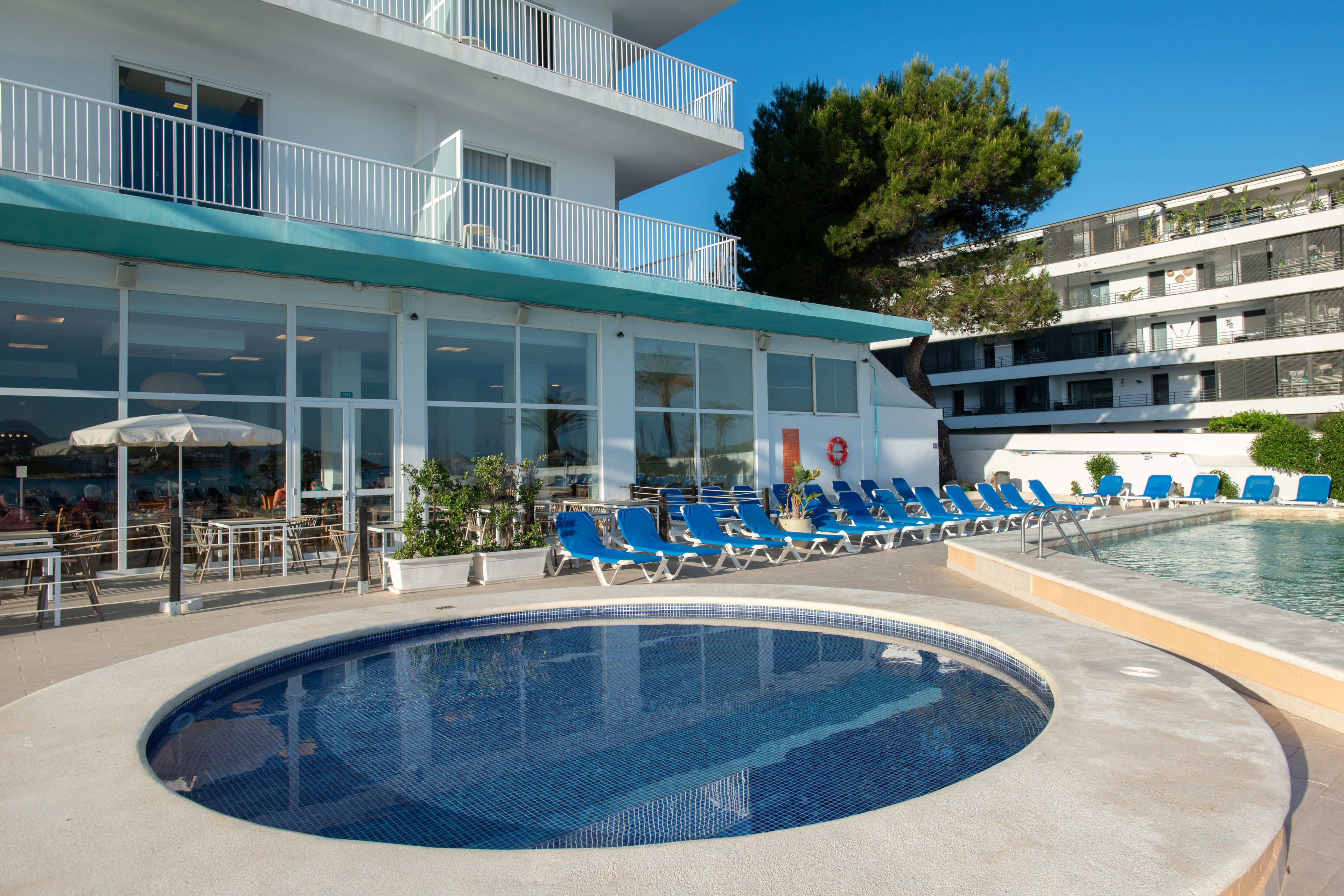 An outdoor hotel pool area features a small circular pool in the foreground and a larger rectangular swimming pool beside it. A long row of blue sun loungers lines the pool deck, facing the water and the modern hotel building. Large glass windows reveal an indoor dining area, while balconies and a green awning overlook the terrace. The scene is brightly lit under a clear blue sky, suggesting a warm, relaxing vacation atmosphere.