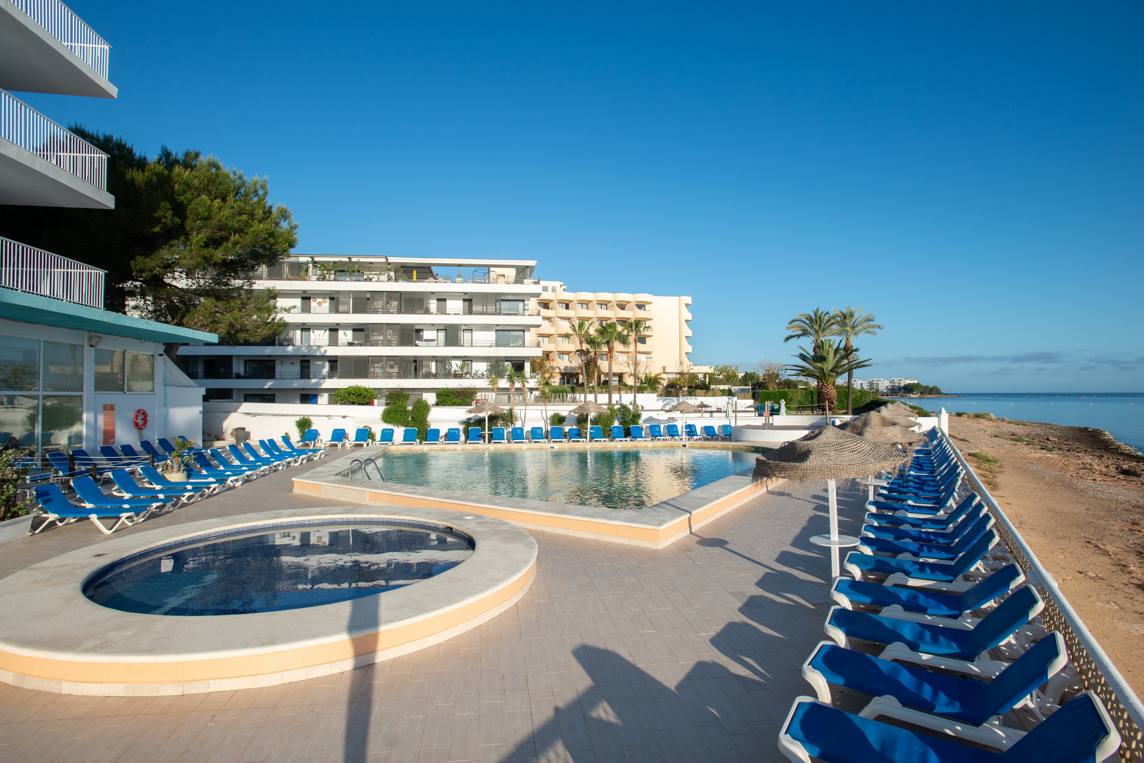 A circular children’s pool and a larger main pool sit beside a modern seaside hotel. Long rows of empty blue sun loungers line the tiled pool deck, extending toward the sandy shoreline. The calm sea and clear blue sky create a peaceful coastal atmosphere. Palm trees and multi-story resort buildings frame the relaxed vacation setting.