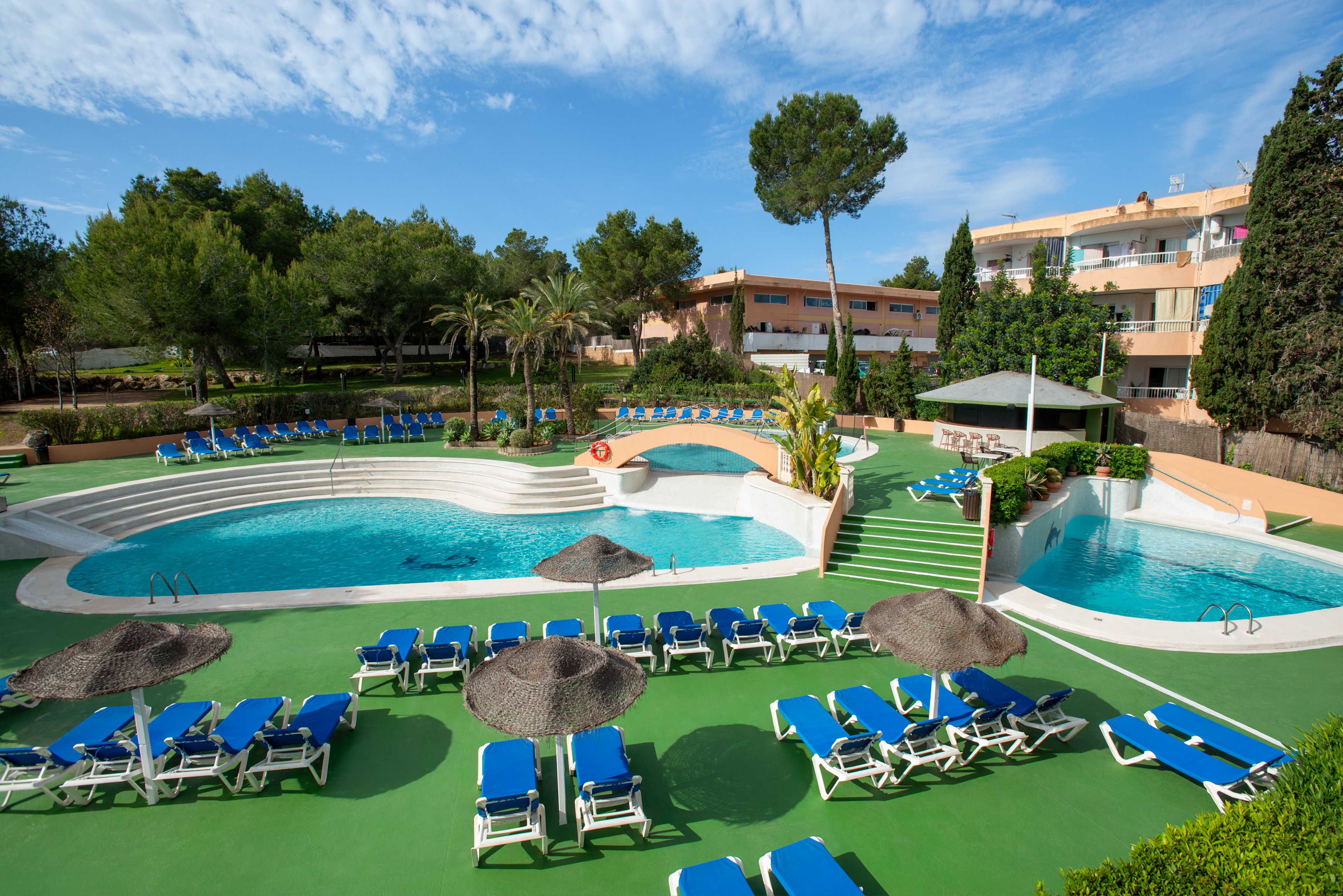 Outdoor hotel swimming pools are surrounded by numerous blue sun loungers and parasols. The scene shows a sunny day with clear skies and lush green trees around the resort buildings. A small bridge crosses part of the pool, and steps lead up to a shaded terrace area. The overall setting suggests a relaxing vacation atmosphere at a Mediterranean-style resort.