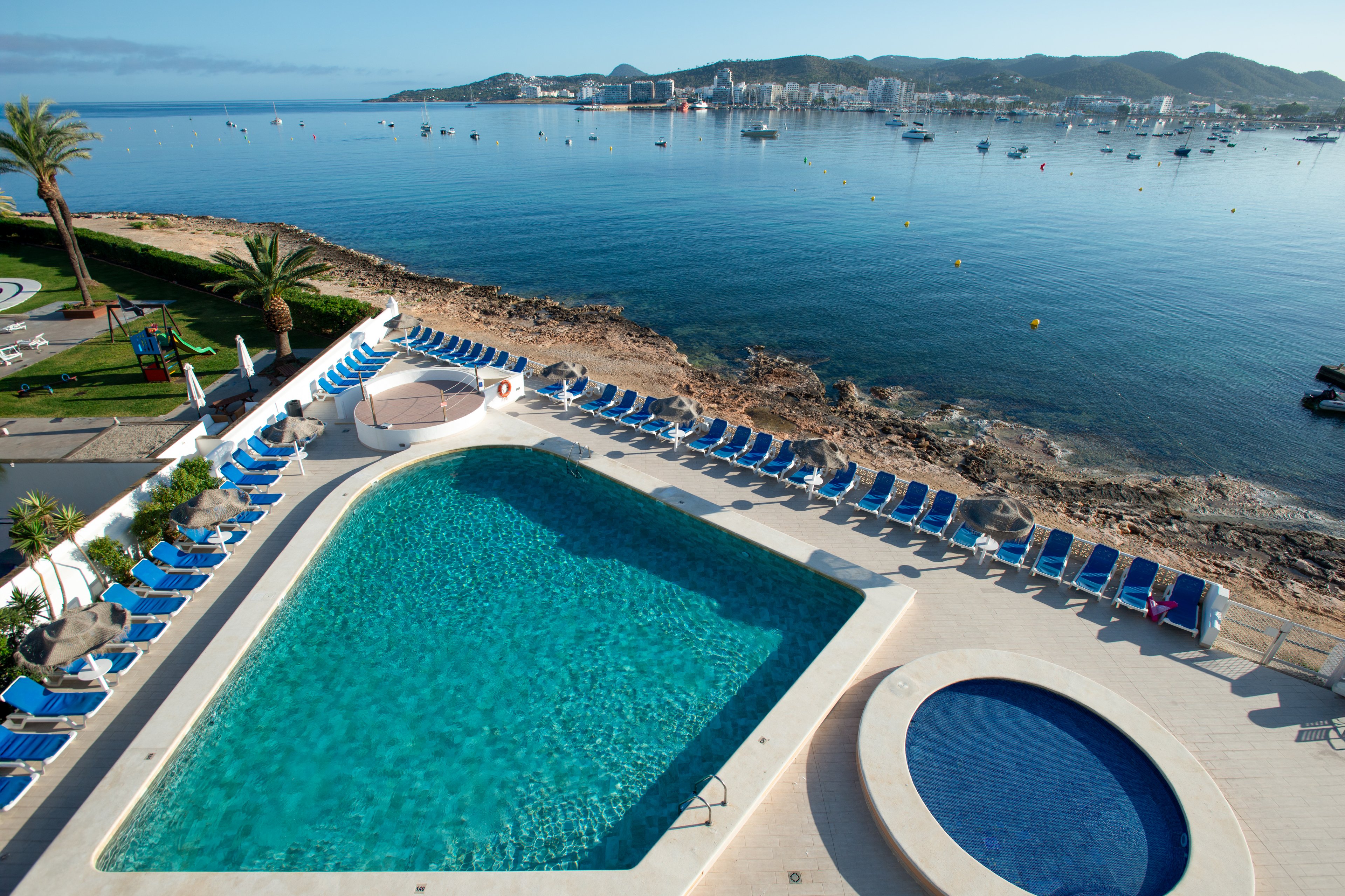 A large rectangular swimming pool sits beside a smaller circular pool on a terrace overlooking a tranquil bay. Numerous blue sun loungers line the pool deck, facing the water and rocky shoreline. The sea is calm with small boats anchored in the distance and low hills on the horizon. The scene suggests a relaxing coastal resort environment under clear skies.